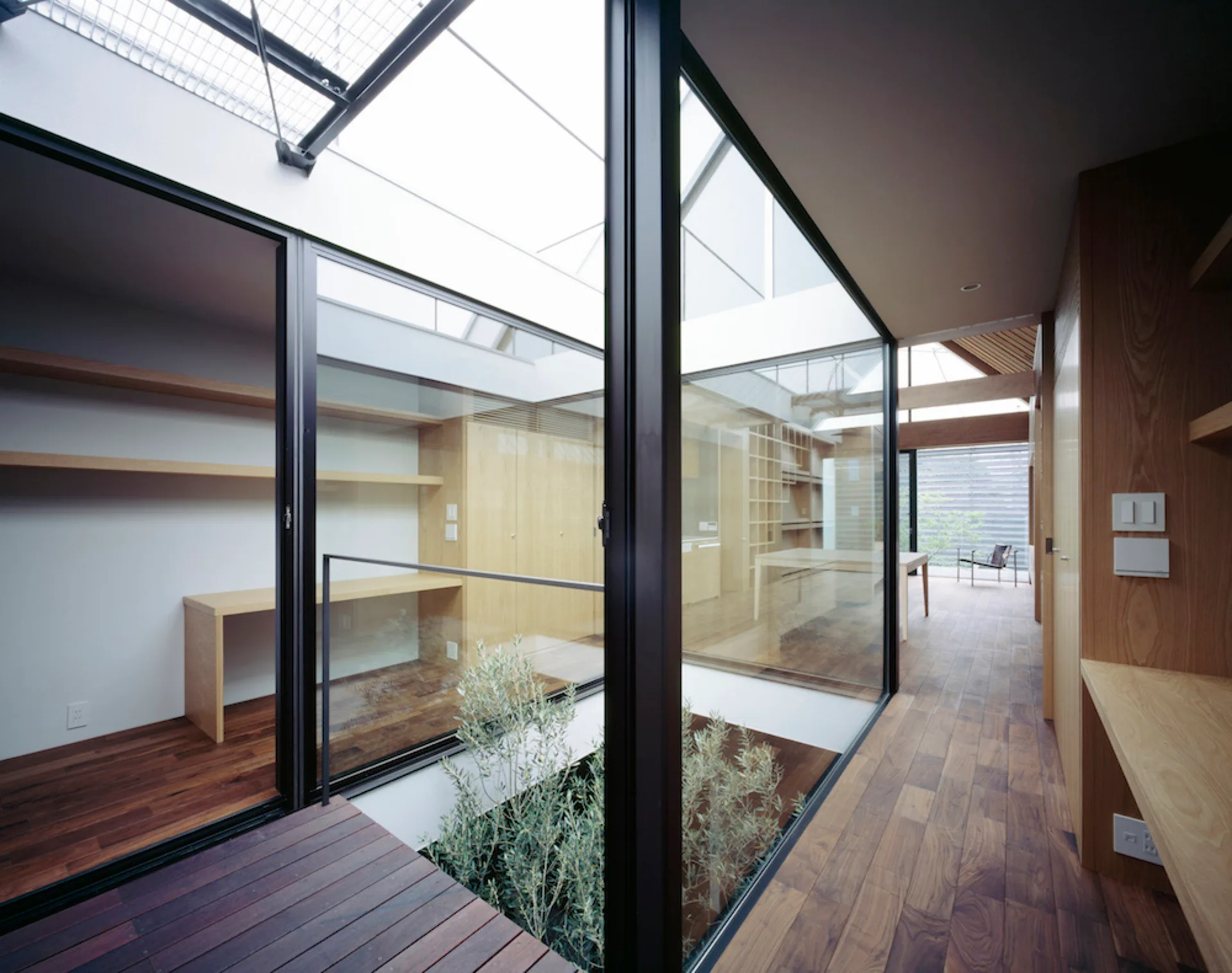 A modern home interior features a central glass-enclosed courtyard with a small plant, surrounded by wood floors and built-in desks.