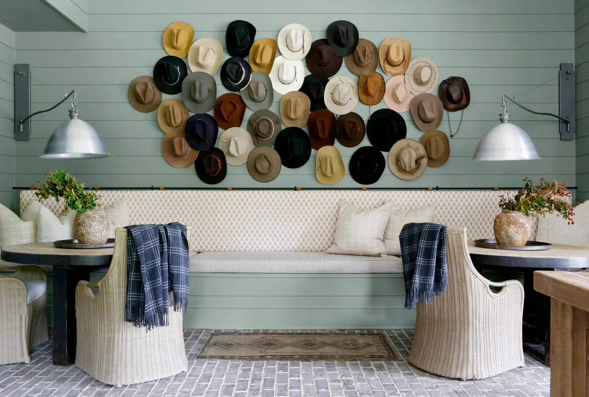 A sage green paneled dining nook features a built-in banquette, two wicker chairs, round tables, and a gallery wall of cowboy hats.