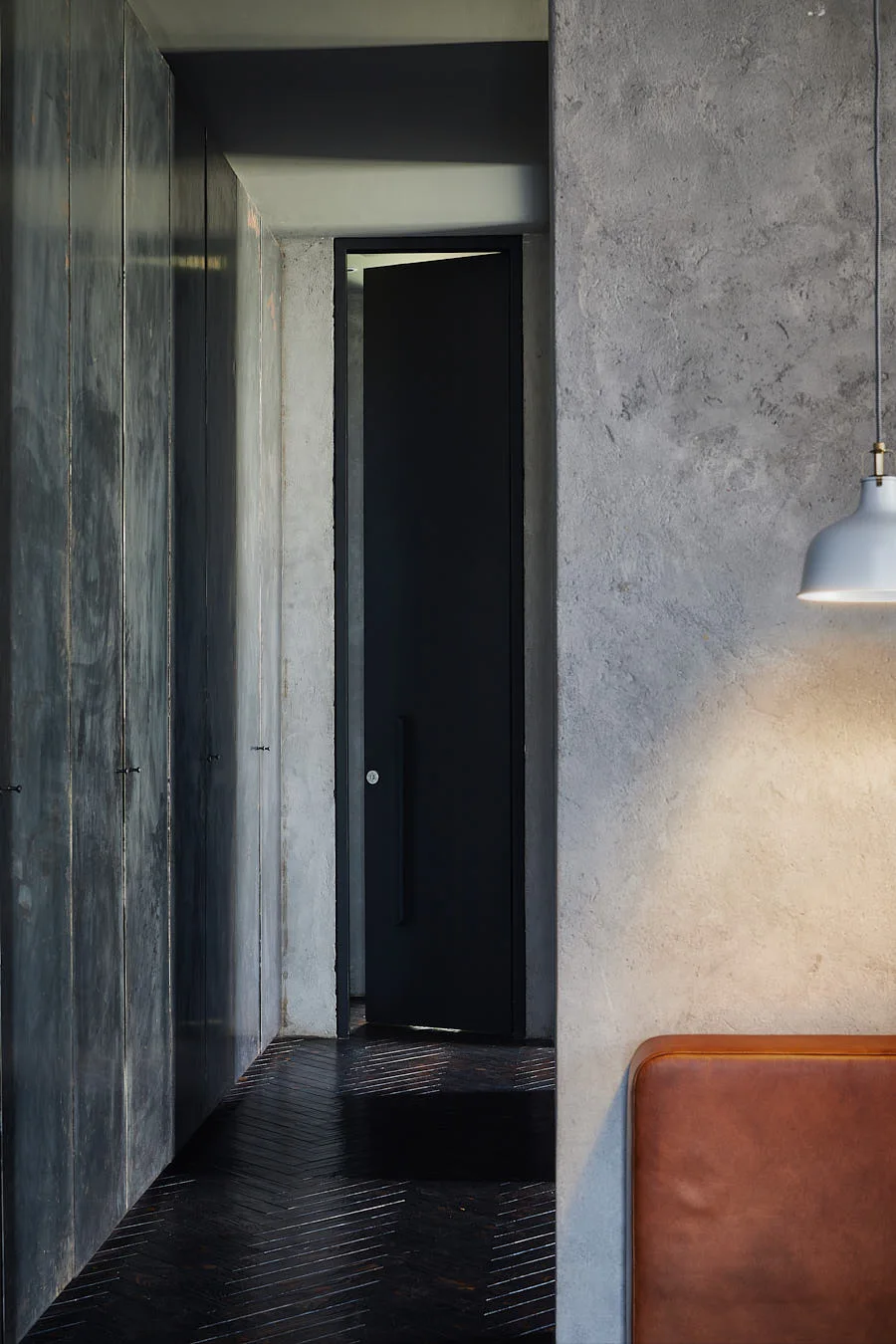 A minimalist hallway featuring textured concrete walls, built-in dark cabinetry, dark herringbone wood flooring, and a black door.