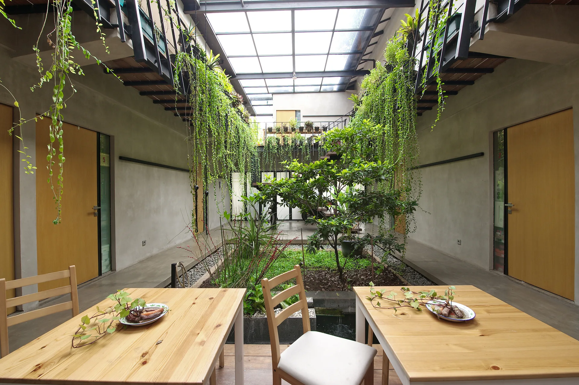Two light wood dining tables sit in a concrete courtyard under a large glass skylight featuring a central garden and hanging green vines.
