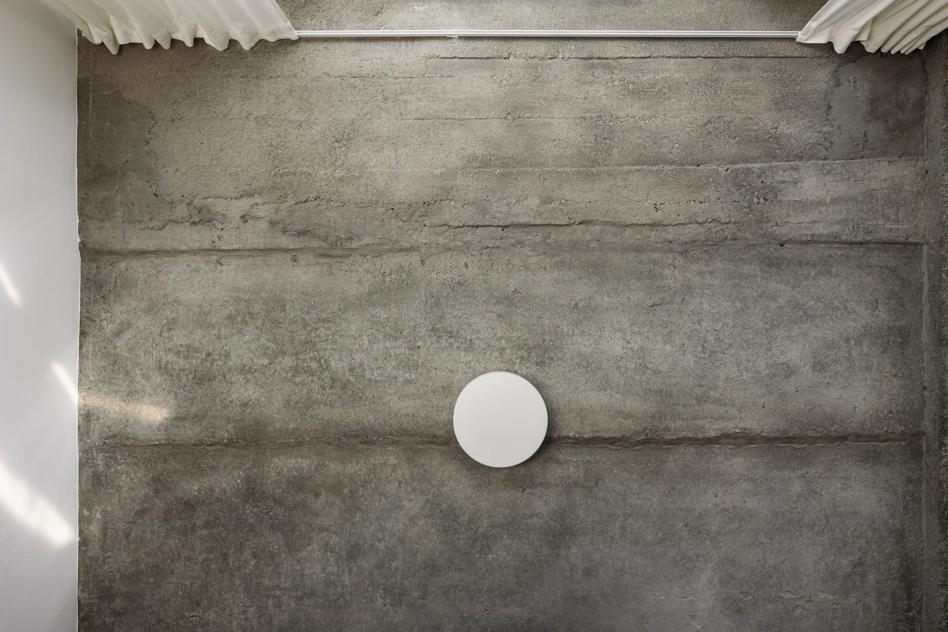 A circular white light fixture is mounted on a textured grey concrete wall, with white pleated curtains visible at the top edge.