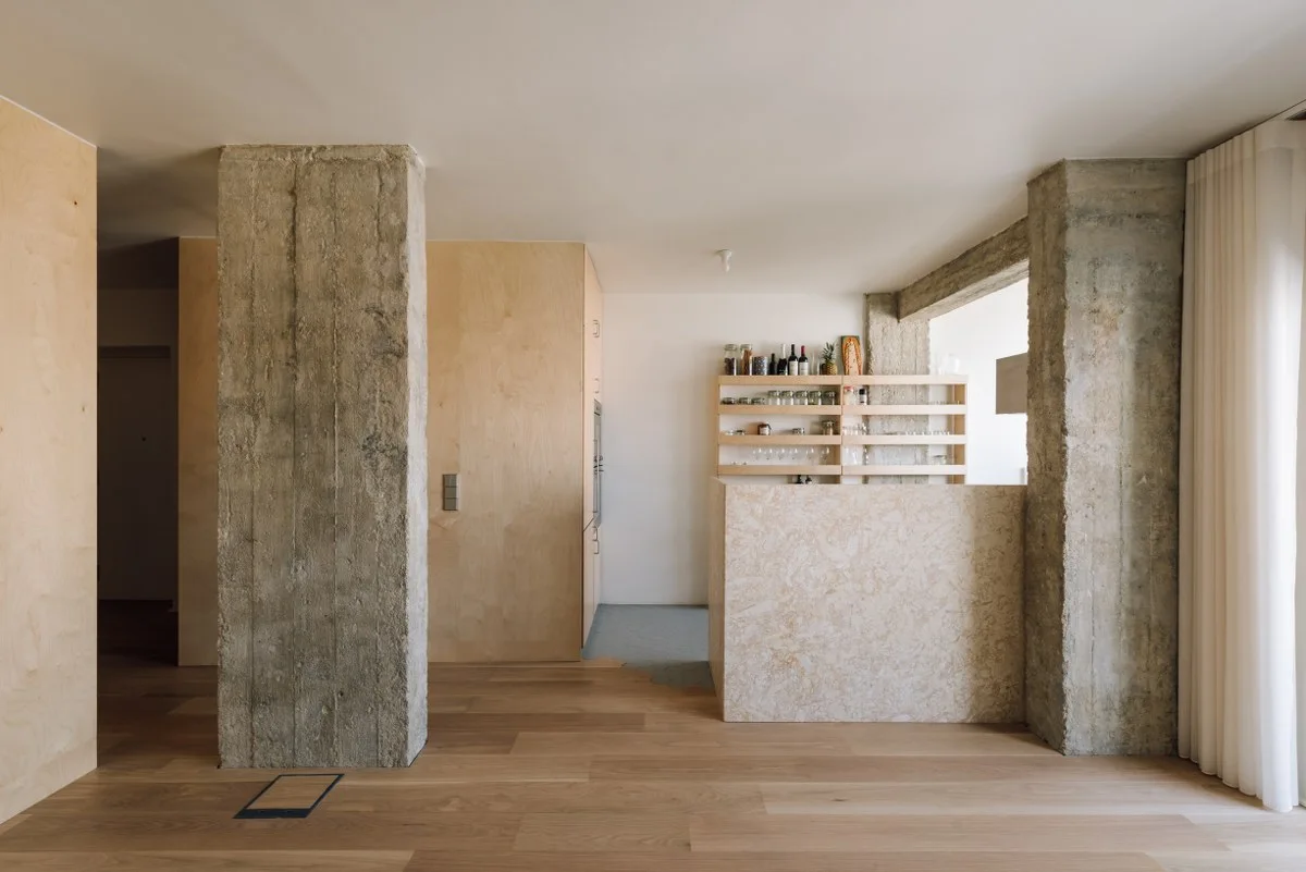 A modern residential interior features two rough concrete columns, plywood walls, a textured stone-like kitchen island, and open wood shelving.