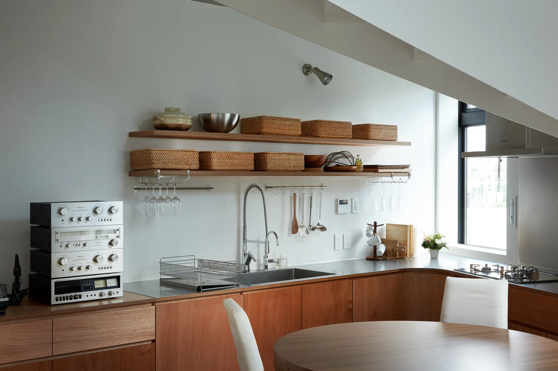 A modern kitchen with warm wood cabinets features open floating shelves holding wicker baskets beneath a white sloped ceiling.