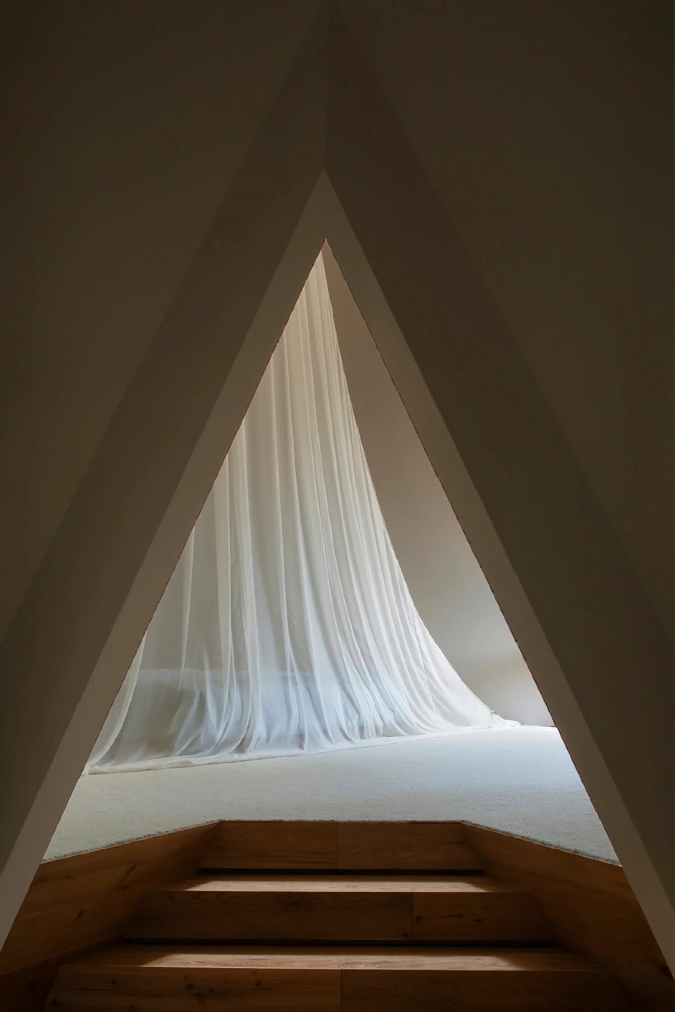 A triangular architectural opening frames a raised minimalist bedroom featuring wooden steps, white carpeting, and a bed draped in a sheer white canopy.