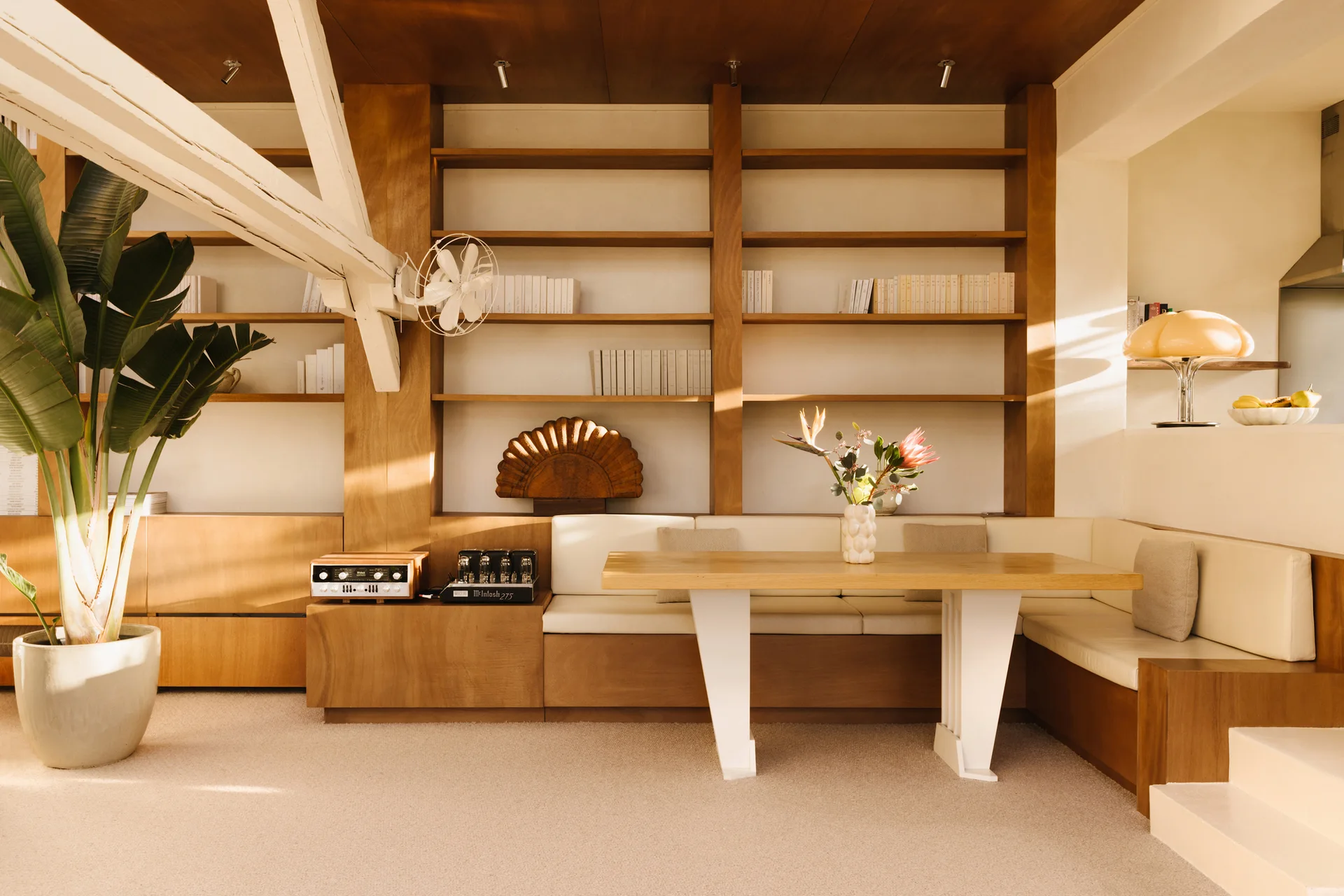 A warm wood dining area featuring a built-in banquette with cream cushions, integrated wall shelving, a light wood table, and a large floor plant.