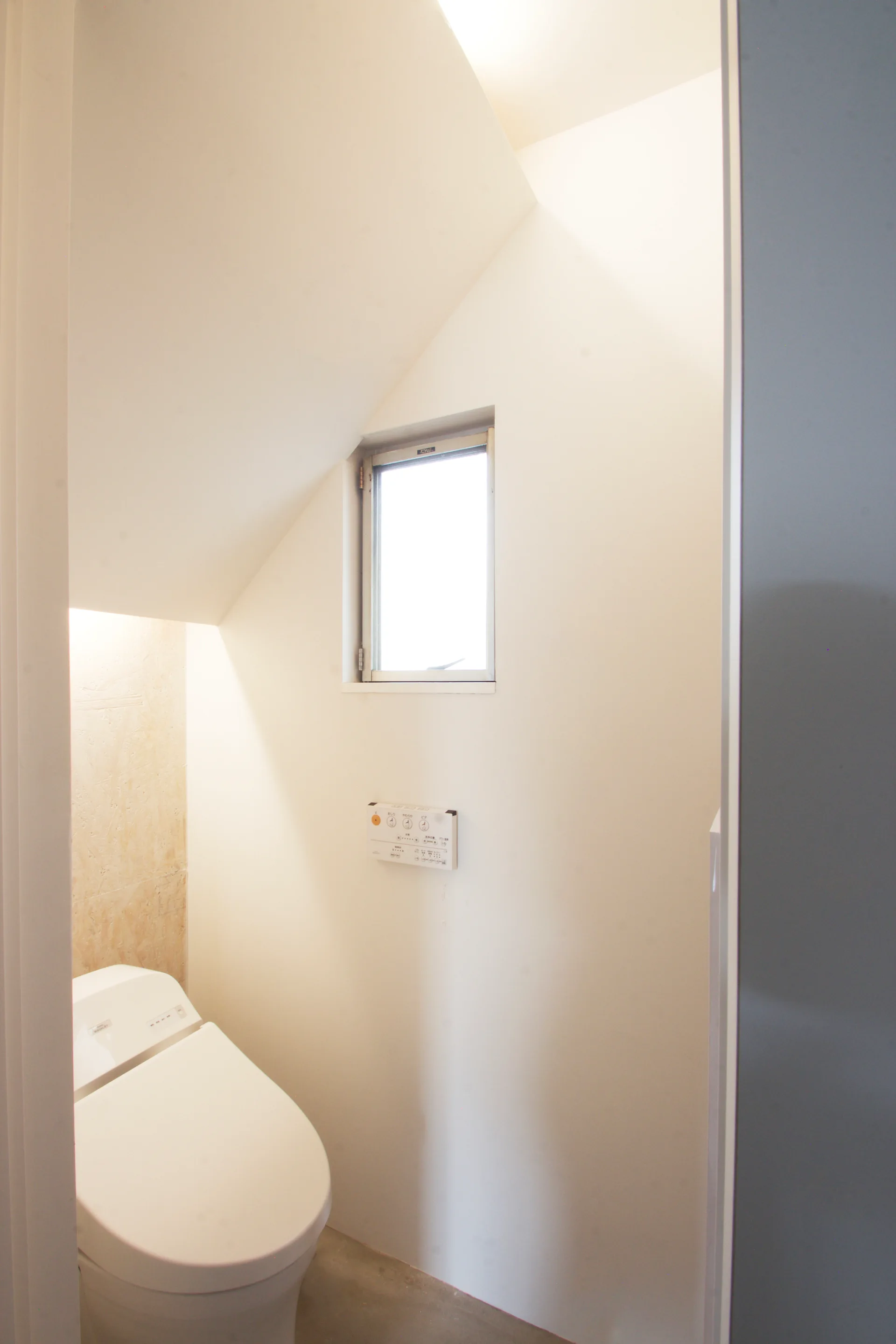 A minimalist white powder room under a sloped ceiling features a modern toilet, a small rectangular window, and a light wood chip OSB accent wall.