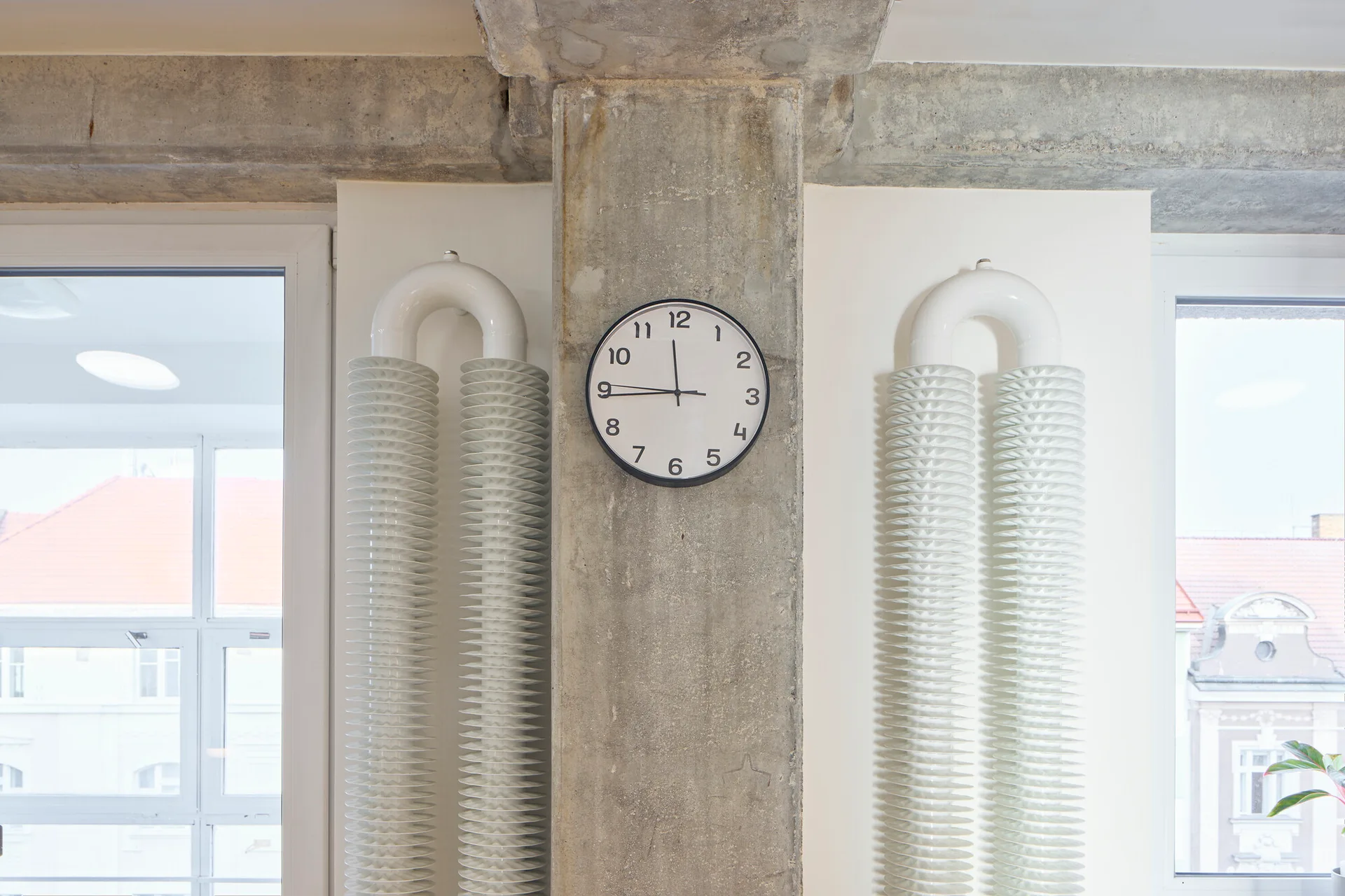 An analog clock hangs on an exposed concrete column between two white coiled loop radiators on a white wall flanked by windows.