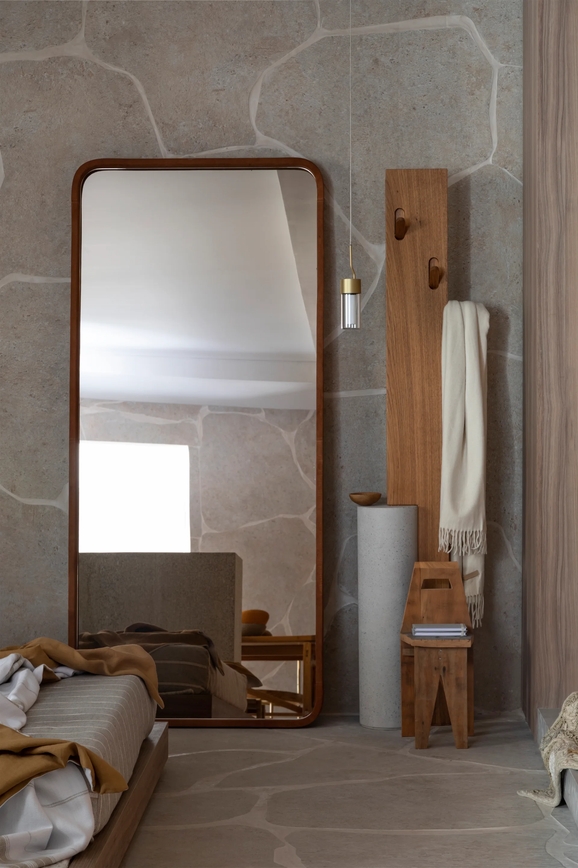 A tall wood-framed floor mirror leans against a beige stone-clad wall next to a wooden coat rack, cylindrical pedestal, and rustic wood stool.