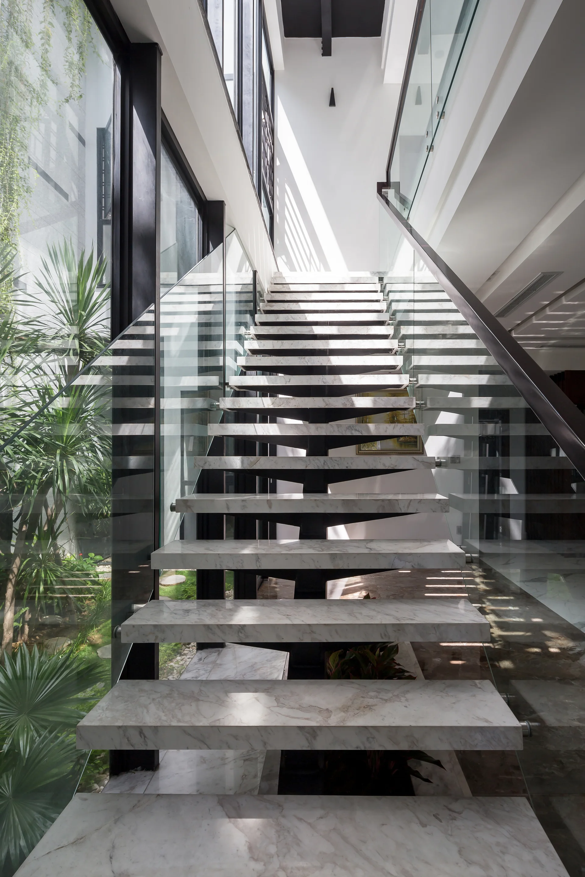 A modern floating staircase with veined white marble treads and glass railings stands next to a tall glass wall overlooking a green courtyard.