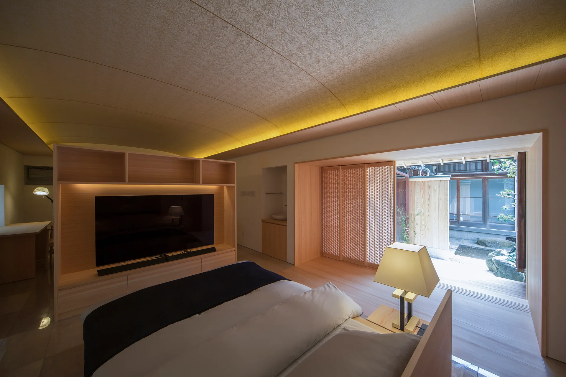 A minimalist bedroom features a low bed, a curved coved ceiling with warm lighting, wood paneling, and sliding screen doors opening to a courtyard.