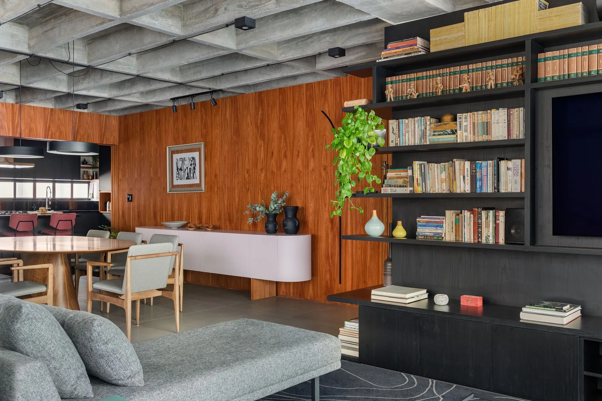 An open-plan living space features an exposed concrete waffle ceiling, vertical wood-paneled walls, a large black bookshelf, and a pink sideboard.