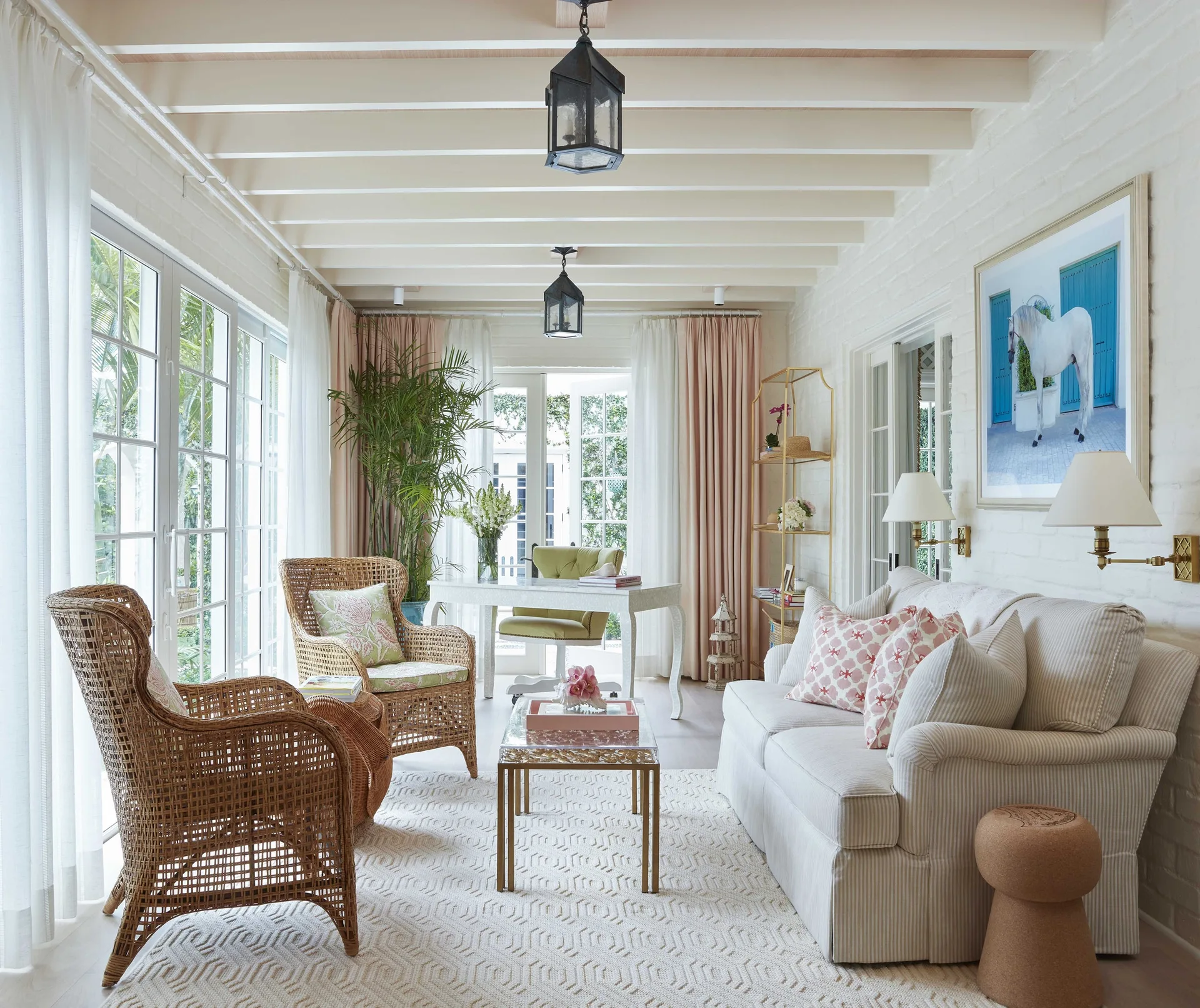A bright sunroom featuring a striped sofa, two wicker armchairs, a gold coffee table, white brick walls, and blush pink drapes.