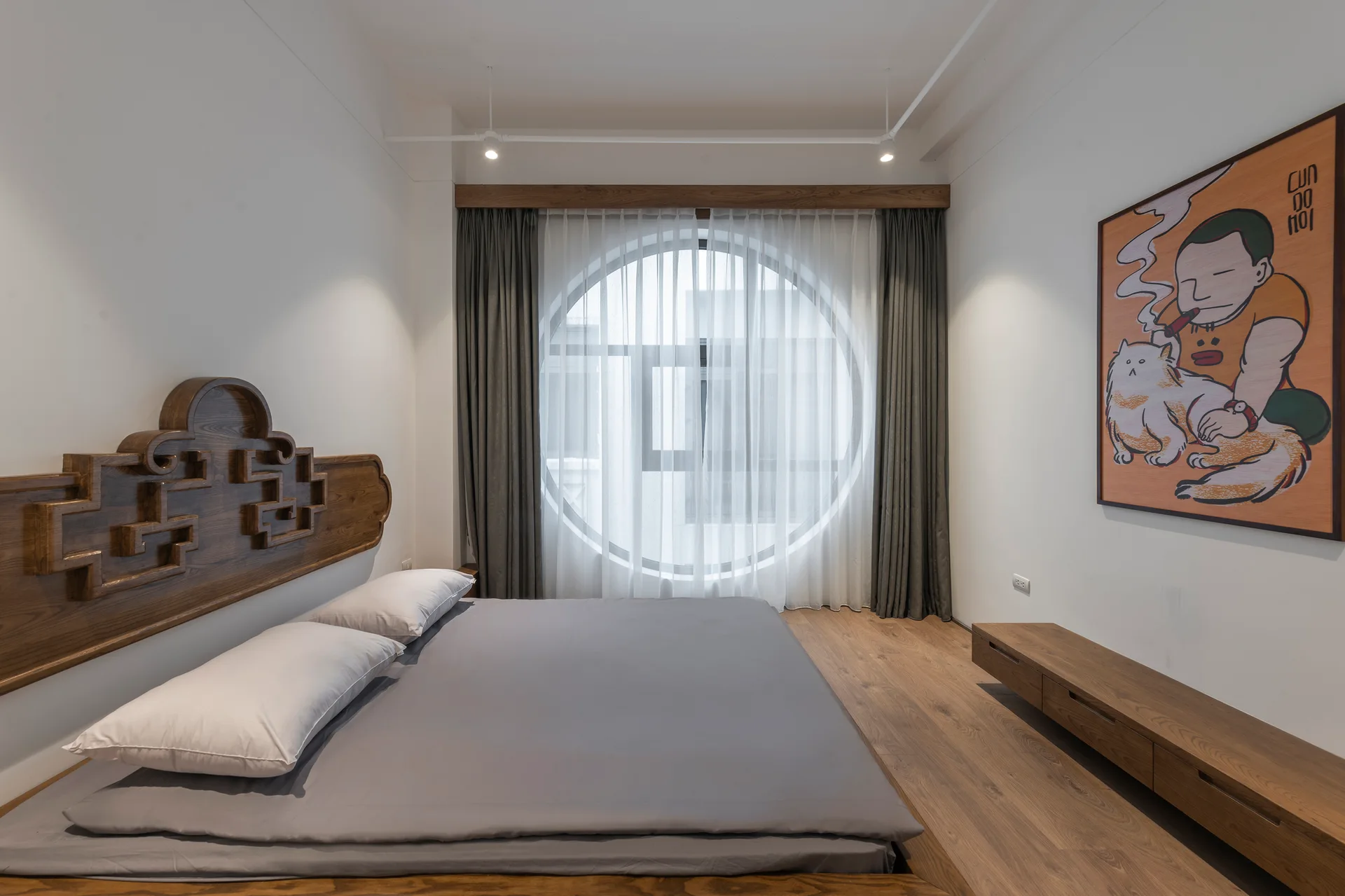 A minimalist bedroom featuring a low platform bed with a carved wooden headboard, white walls, and a large circular window with gray drapes.