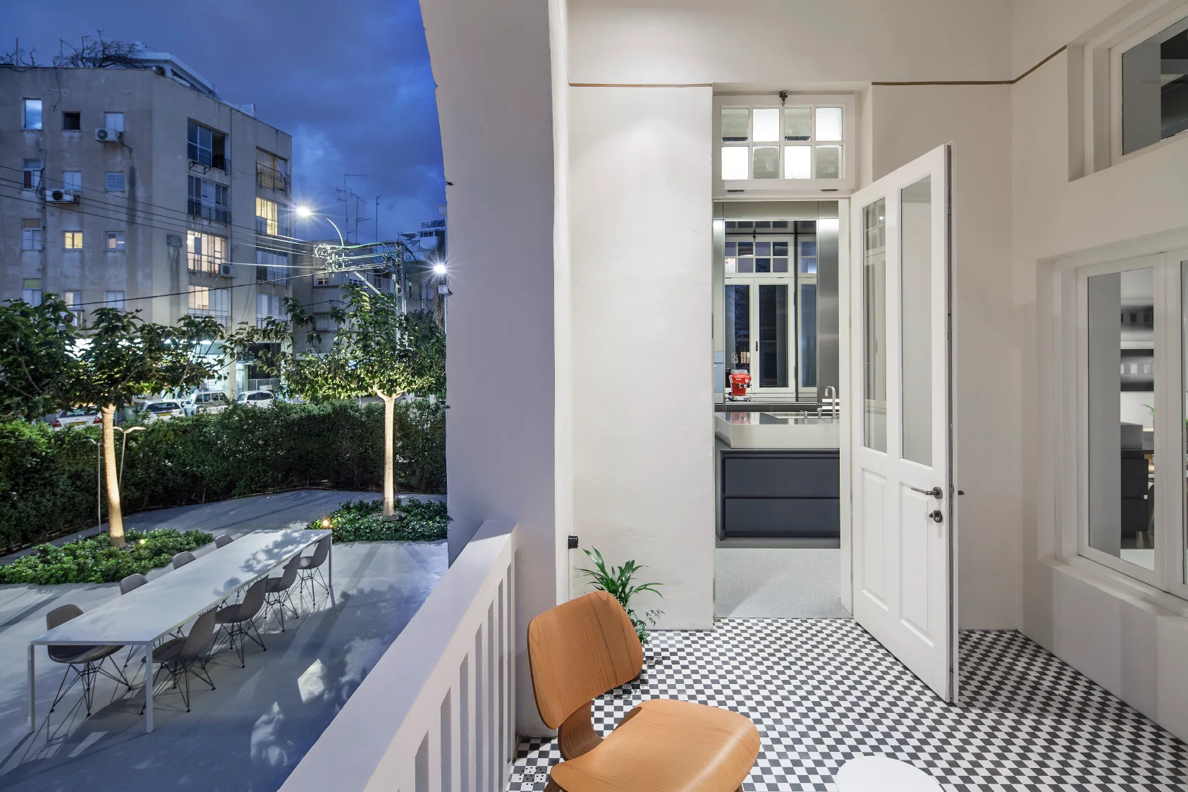 A covered balcony with a black and white checkered tile floor, a wooden chair, and an open door showing a modern kitchen at night.