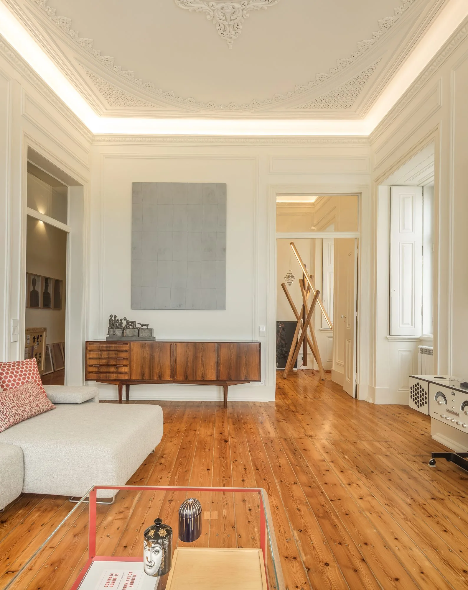 A living room features a mid-century wooden sideboard, a light gray sofa, polished hardwood flooring, and a highly detailed ornate plaster ceiling.
