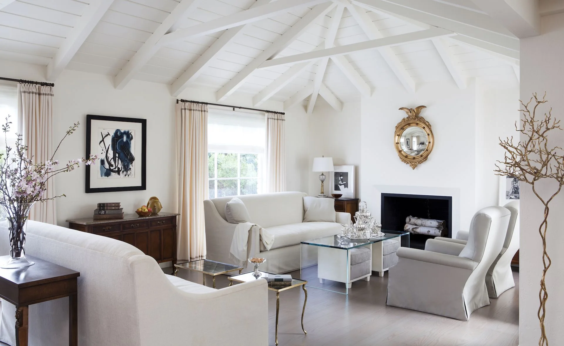 A bright living room features cream-colored sofas, slipcovered armchairs, a central fireplace under a gold mirror, and a vaulted ceiling with white beams.