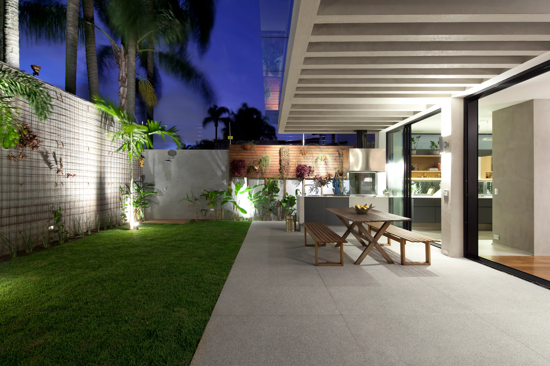 A modern outdoor patio at night features a wooden dining table and benches beneath concrete ceiling beams next to a green lawn.