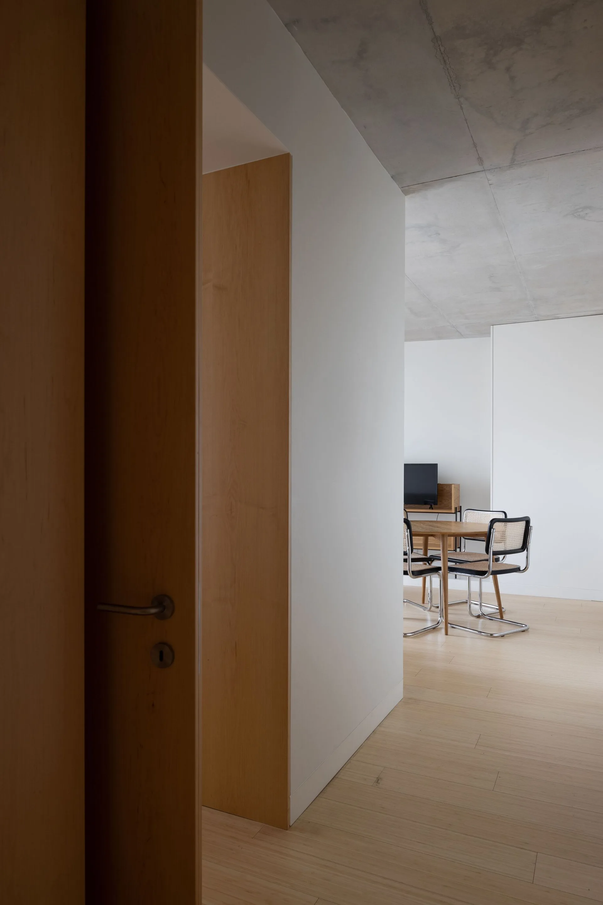 A view through a wood-clad doorway into a minimalist dining room featuring a round wooden table, woven cane chairs, and an exposed concrete ceiling.
