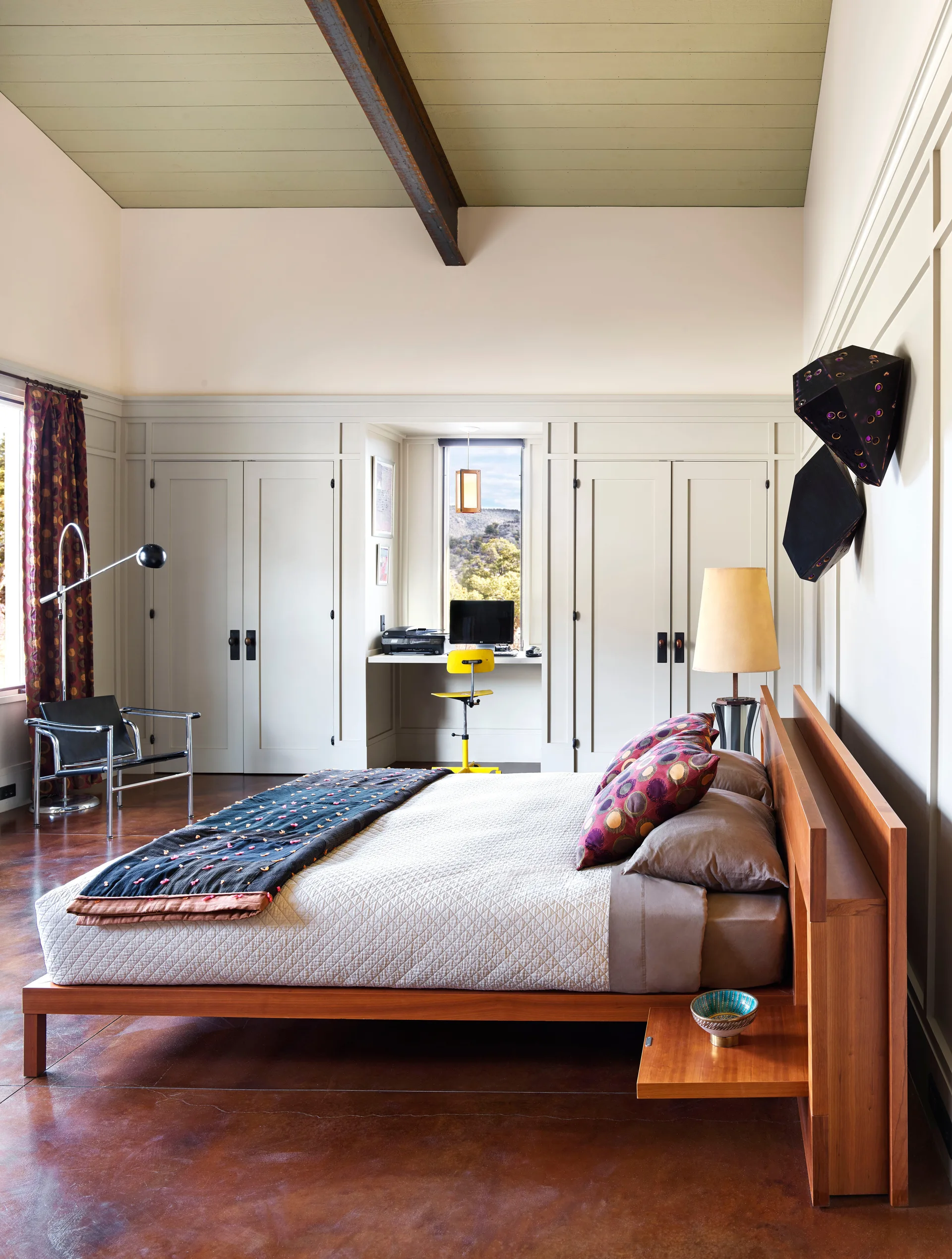 A modern bedroom showing a wooden bed frame, a small built-in desk in an alcove, and floor-to-ceiling cream storage cabinets.