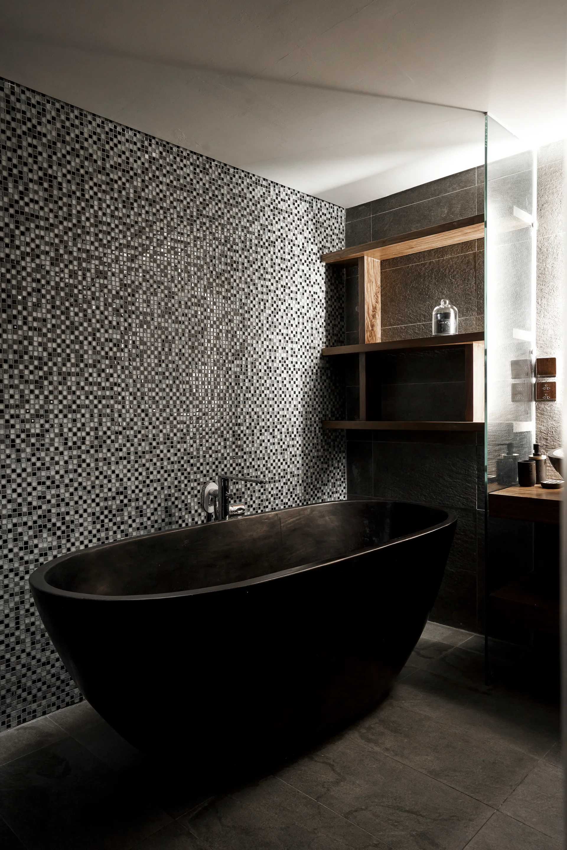 A dark oval freestanding bathtub sits before a black and white mosaic tiled wall, with a wooden shelving unit mounted on an adjacent gray tiled wall.