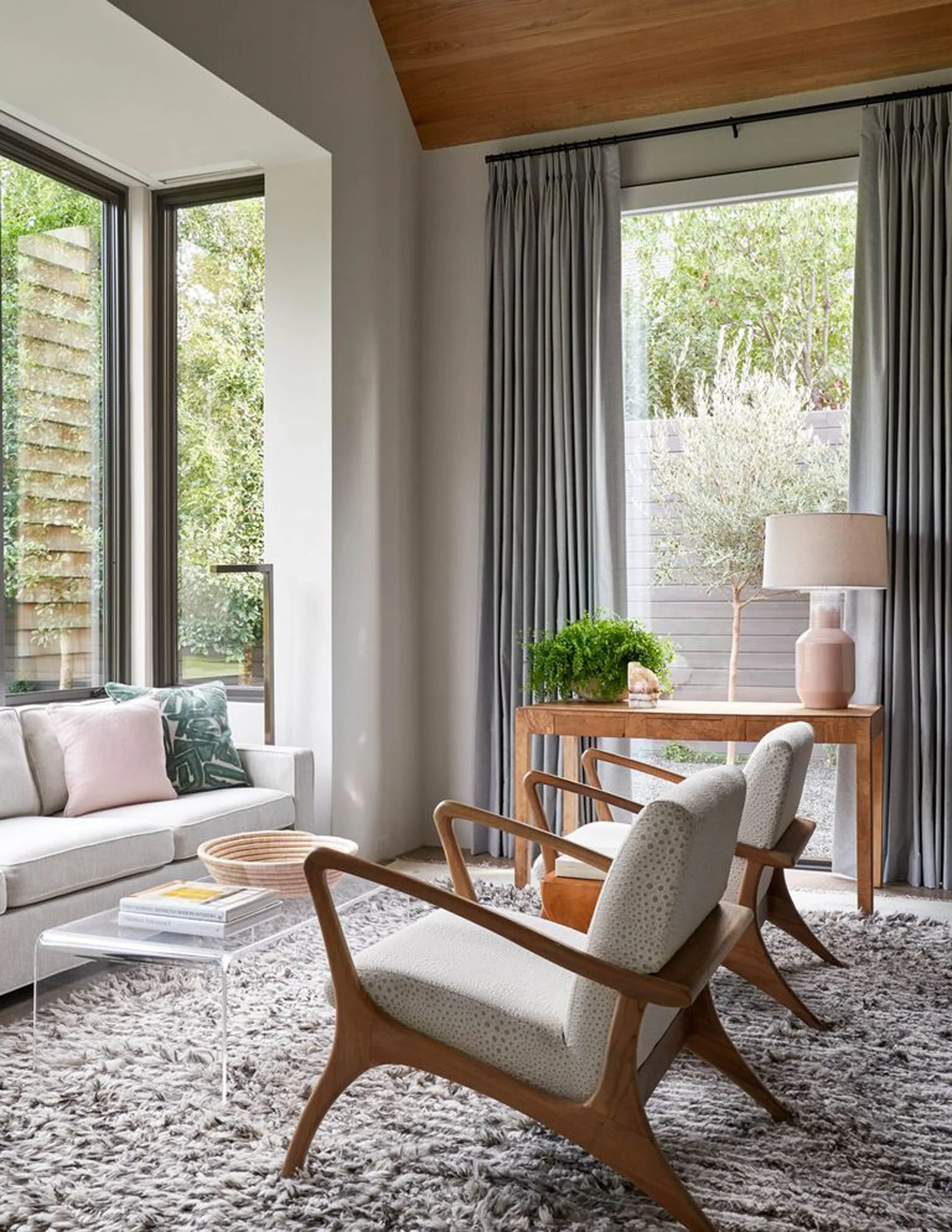 A neutral living room features two mid-century wood armchairs, an acrylic coffee table on a grey shag rug, and a console table by a large window.