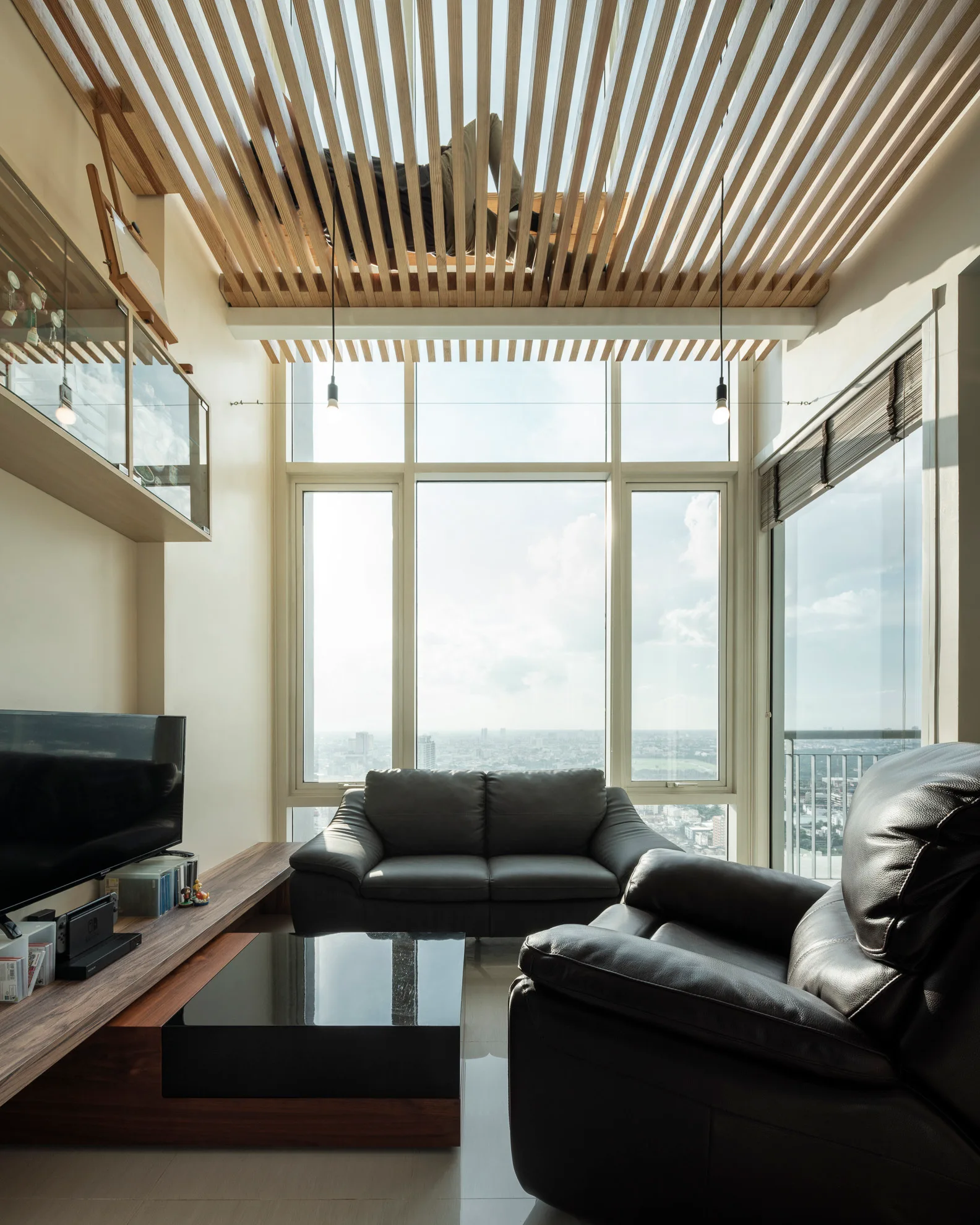 A living room with a black leather sofa, a low wooden media console, large windows showing a skyline, and a slatted wood ceiling.