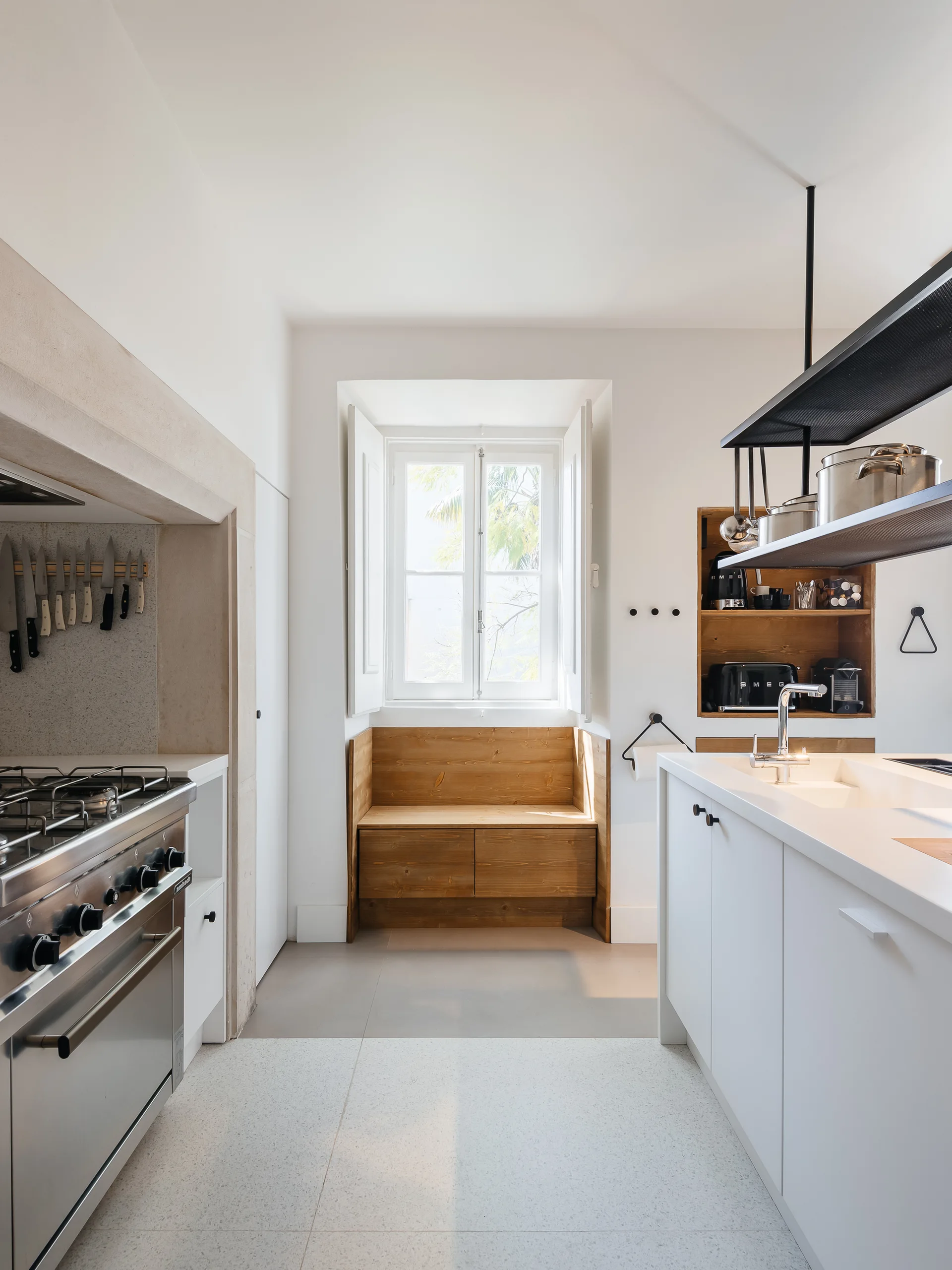 A modern minimalist kitchen features a white central island, a stainless steel range, and a built-in wooden bench in a window nook.
