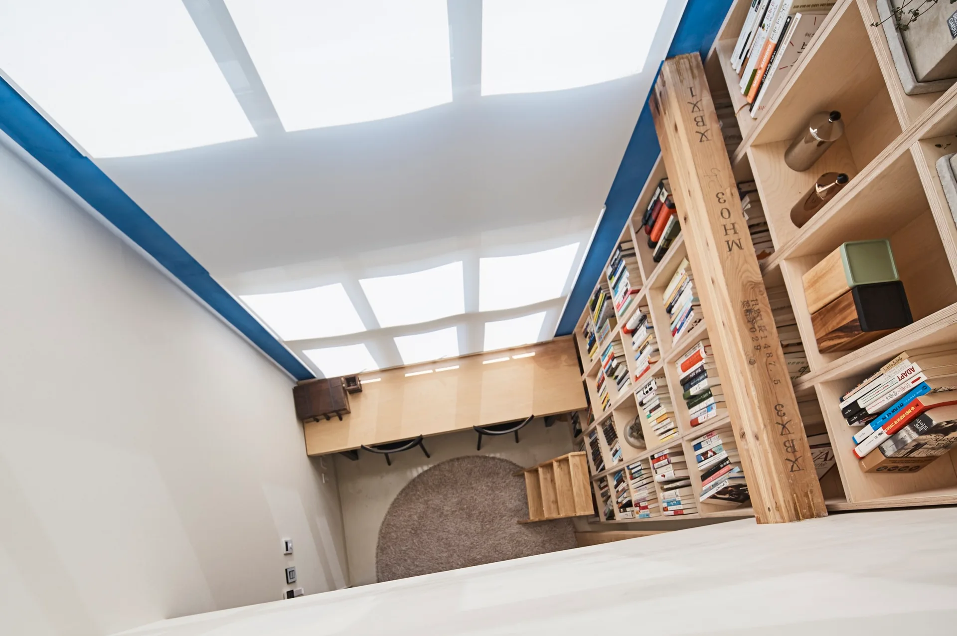A high-angle view of a home library featuring a long built-in wood desk under a grid skylight and tall bookshelves.