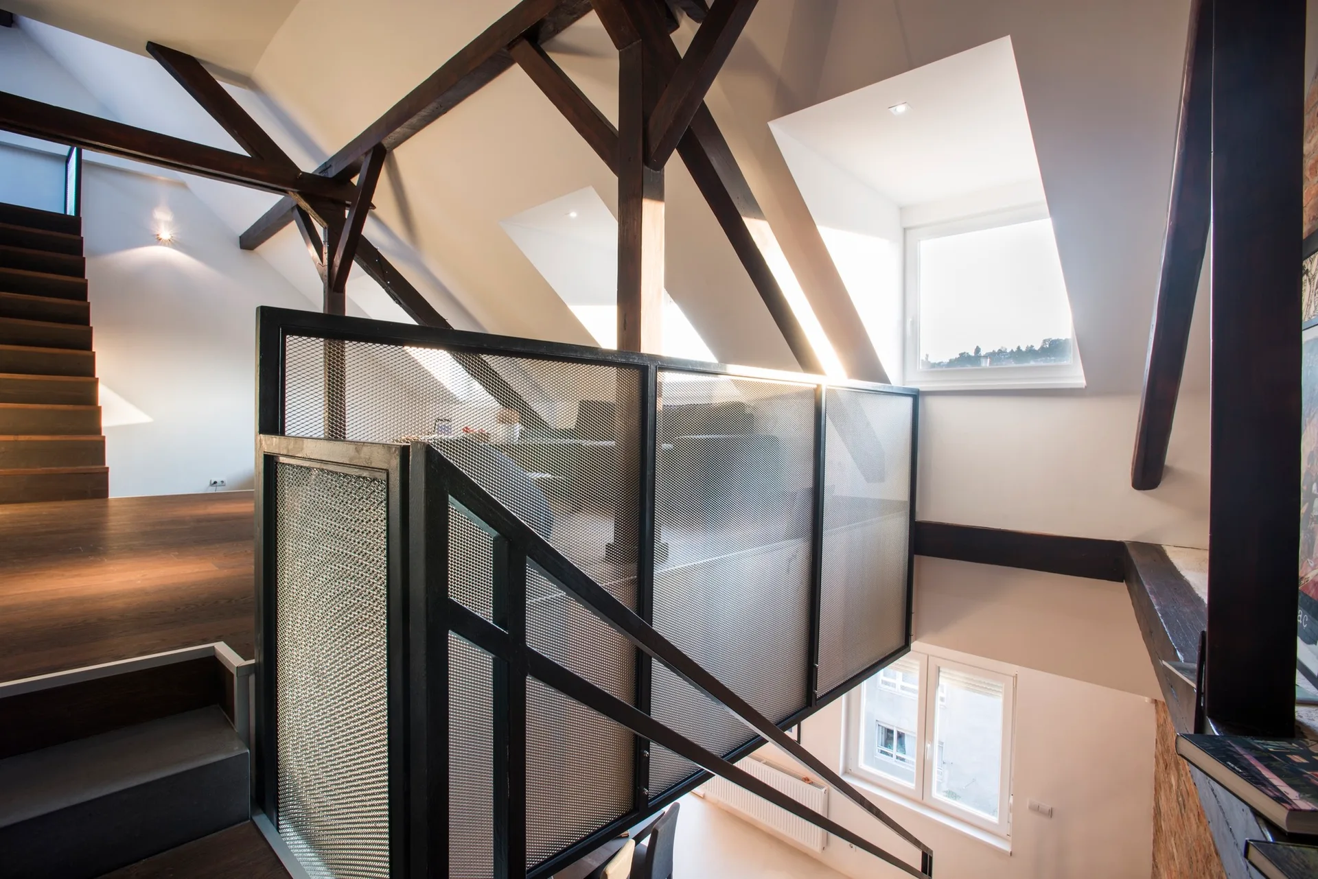 An attic loft features a wooden staircase with black metal mesh railings, set against white walls and dark exposed timber beams.