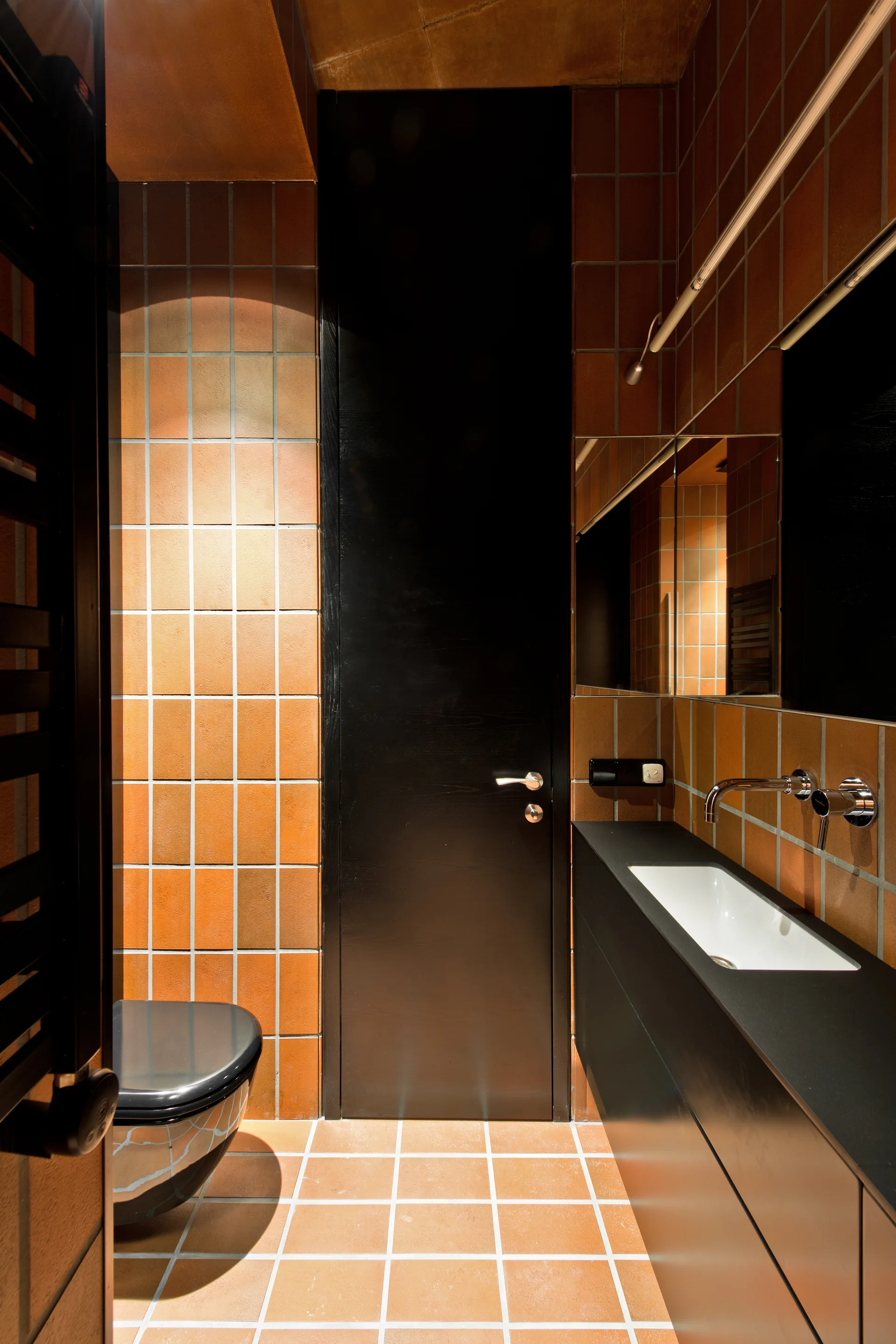 A modern bathroom lined with square terracotta tiles features a black floating vanity with a chrome faucet and a matching black door.