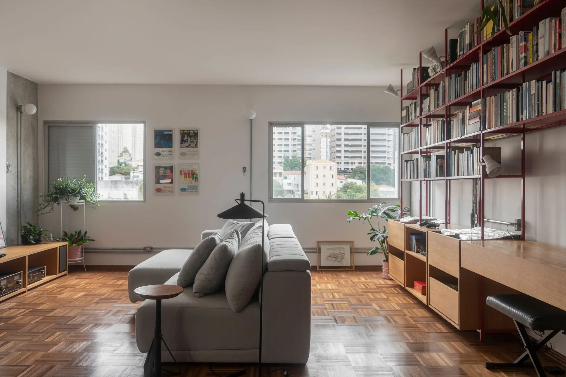 A modern living room features a grey sectional sofa, warm parquet wood flooring, and a large red metal shelving unit filled with books.