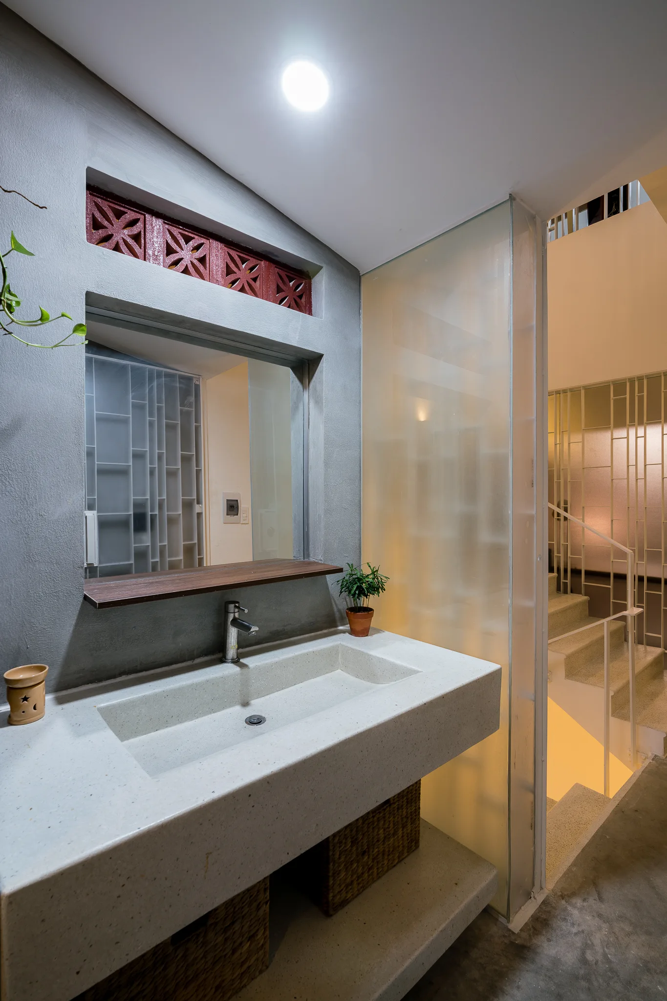 A gray bathroom features a concrete floating vanity with wicker baskets underneath, a mirror, and a frosted glass screen next to a staircase.