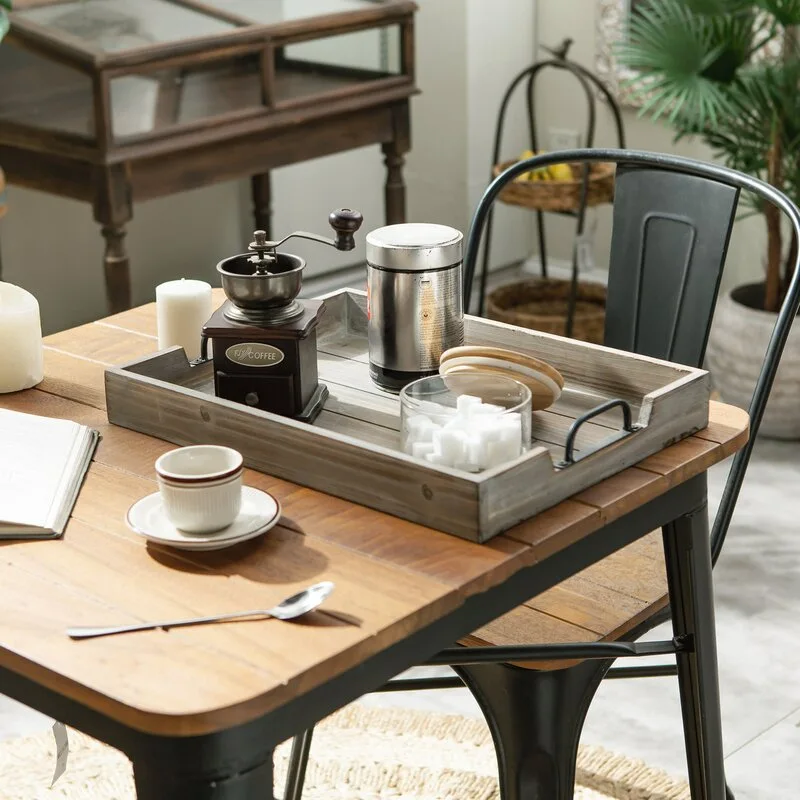 A weathered gray wooden tray holding a manual coffee grinder and sugar bowl sits on a rustic wood and metal dining table.