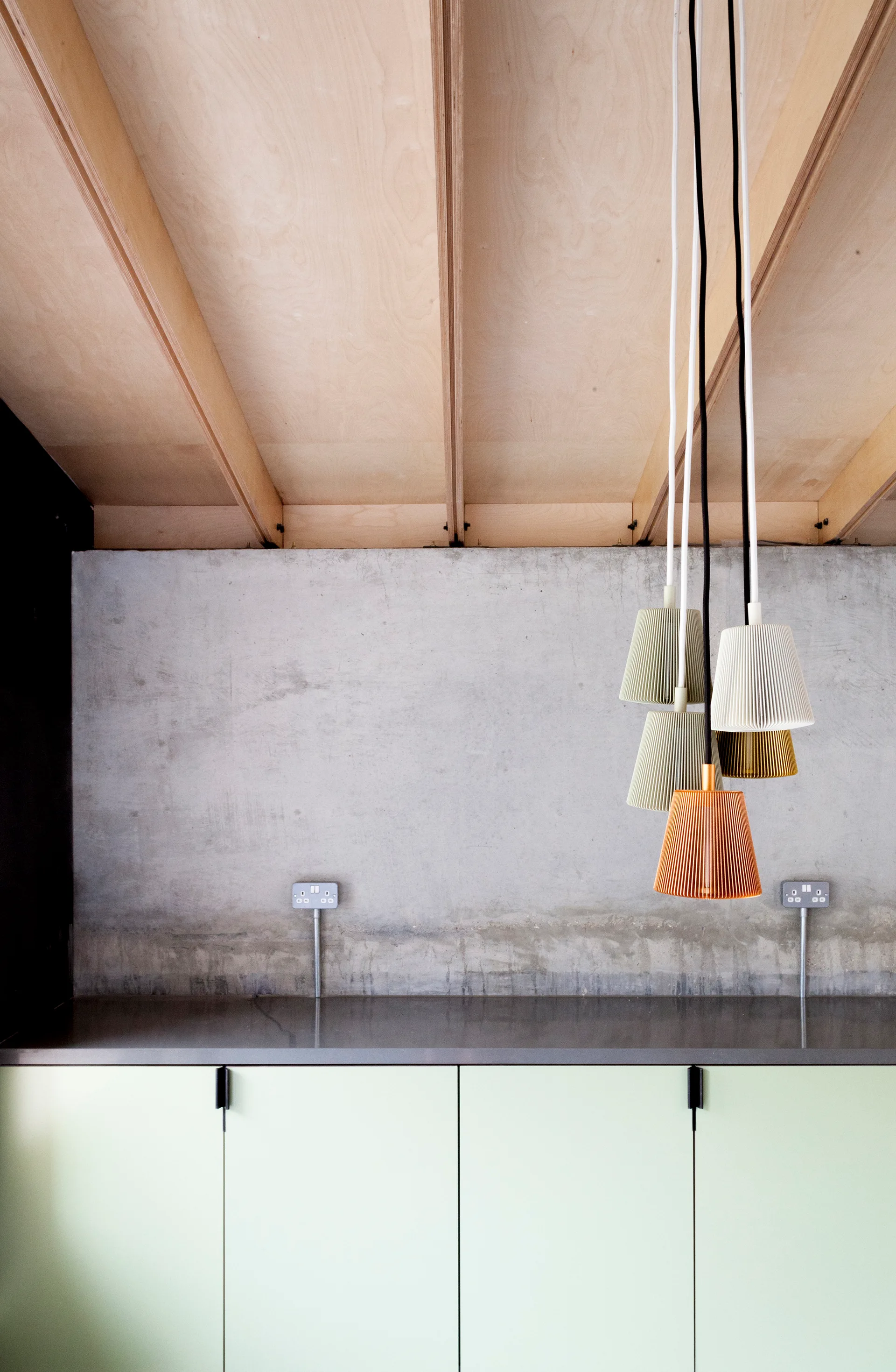 Sage green cabinets stand beneath a raw concrete wall and a cluster of multi-colored ribbed pendant lights hanging from a plywood ceiling.