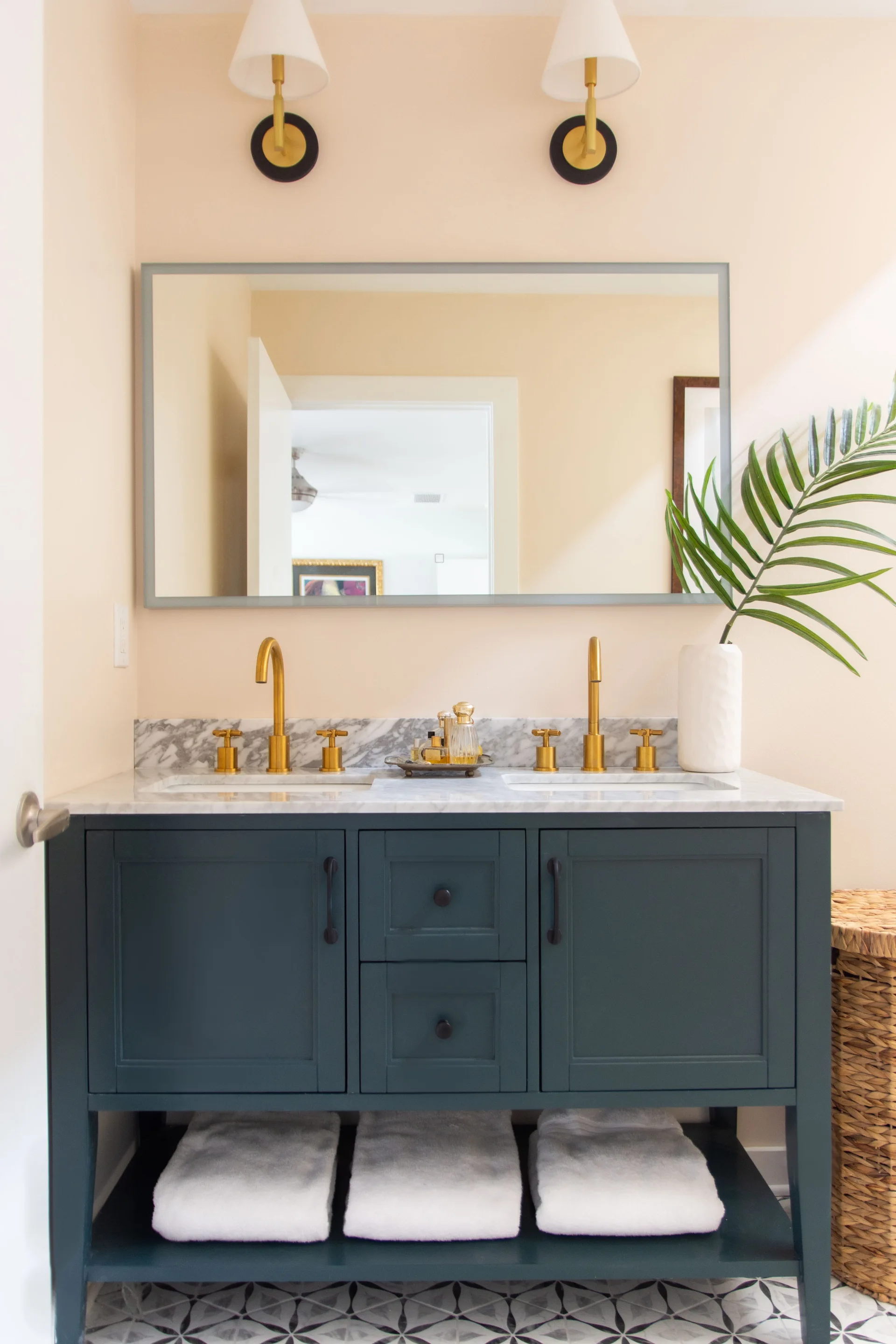 A navy blue double vanity with a white marble countertop, gold faucets, and a large mirror sits against cream walls under two brass sconces.