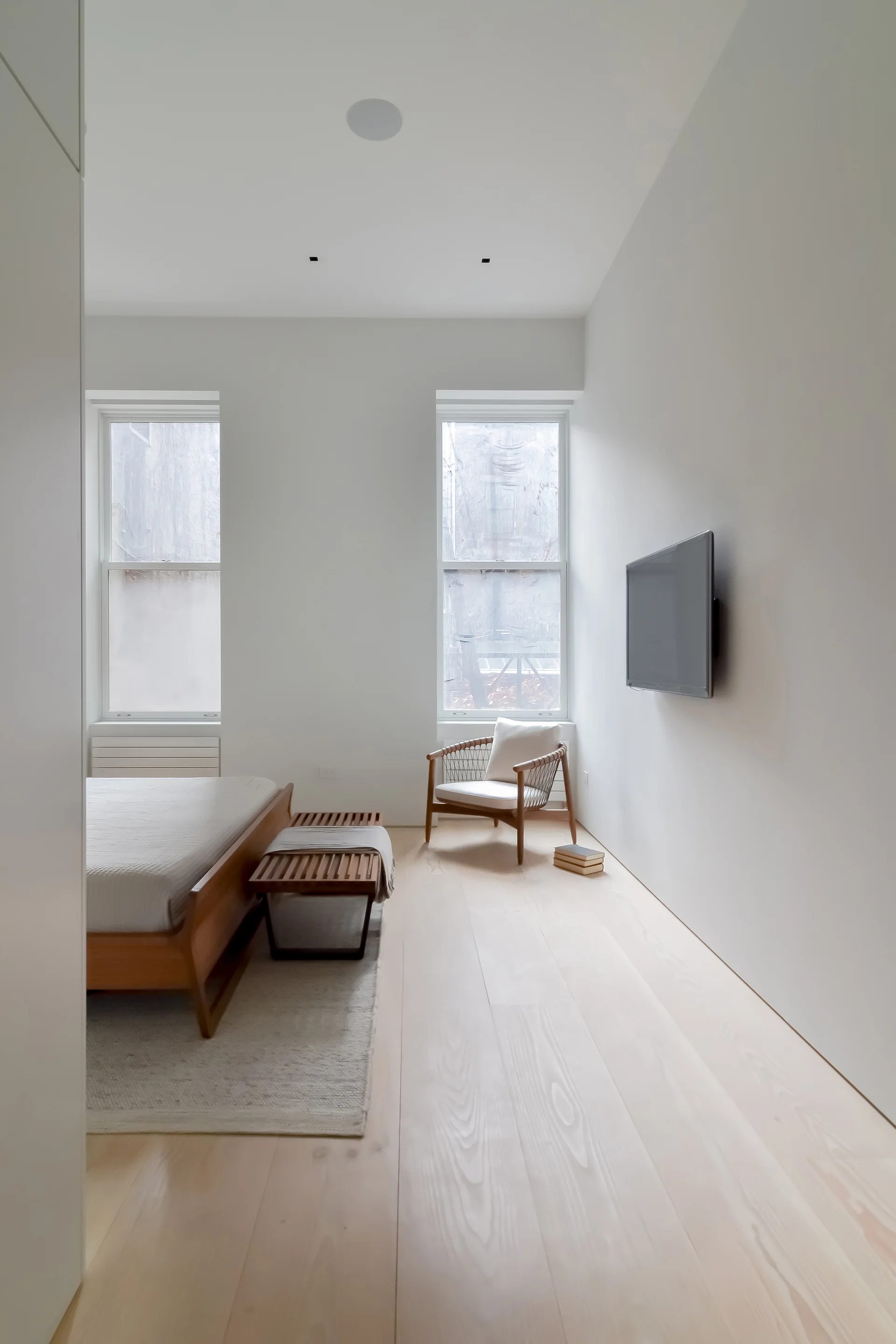 A minimalist bedroom features white walls, wide-plank light wood floors, a platform bed, a slatted wooden bench, and an armchair by the window.