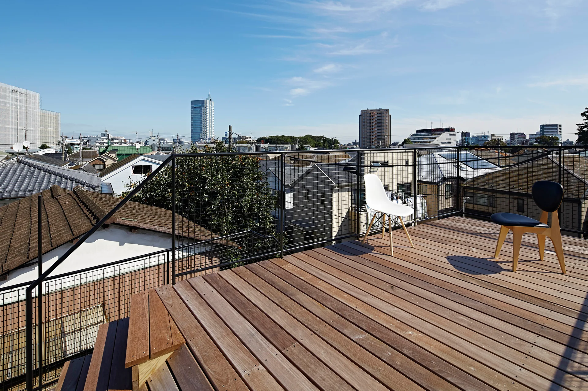 A wooden rooftop terrace features a black grid railing, two minimalist chairs, and views of a city skyline under a clear blue sky.