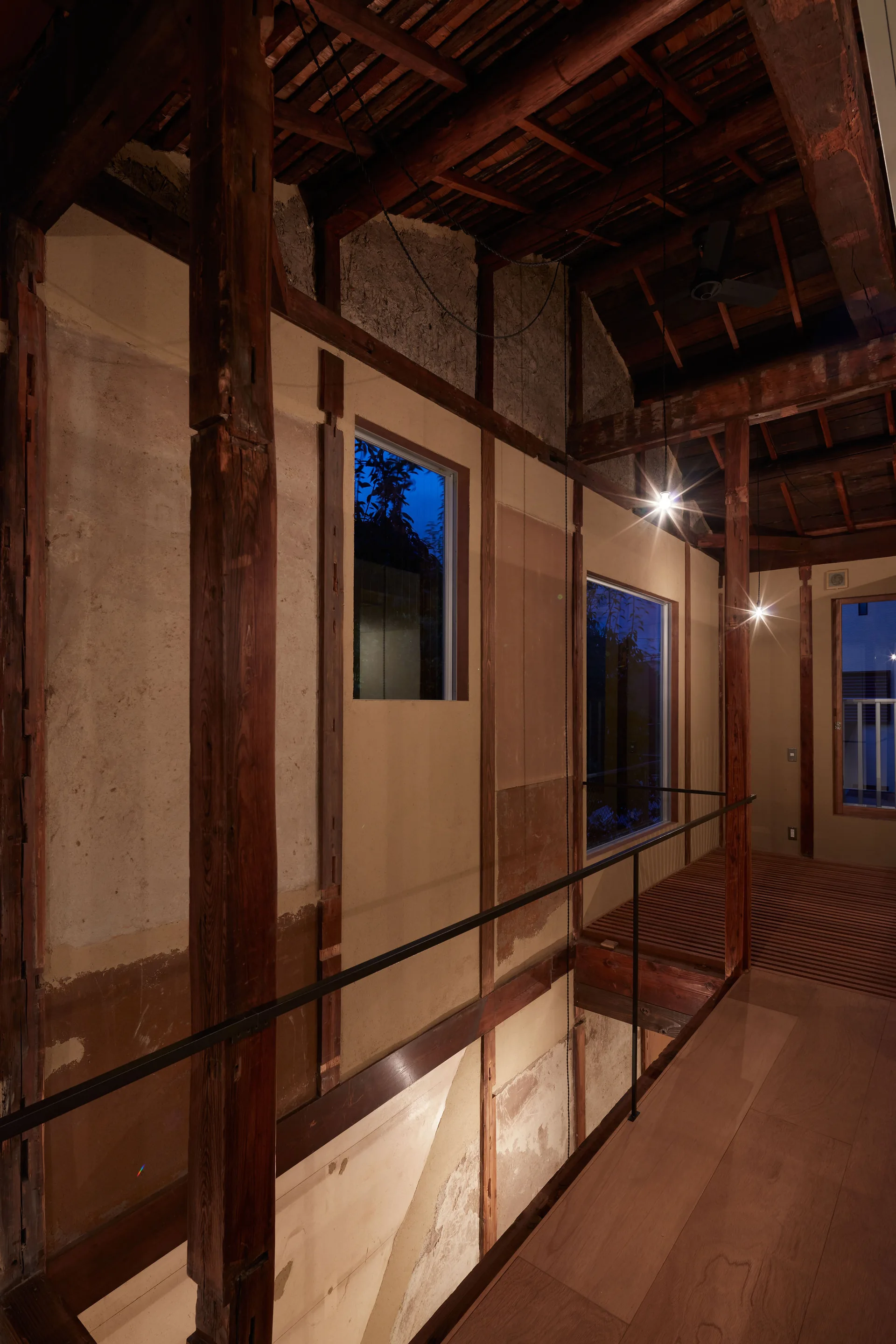 An elevated wooden walkway features exposed timber framing, rustic plaster walls, a thin black metal railing, and small pendant lights.