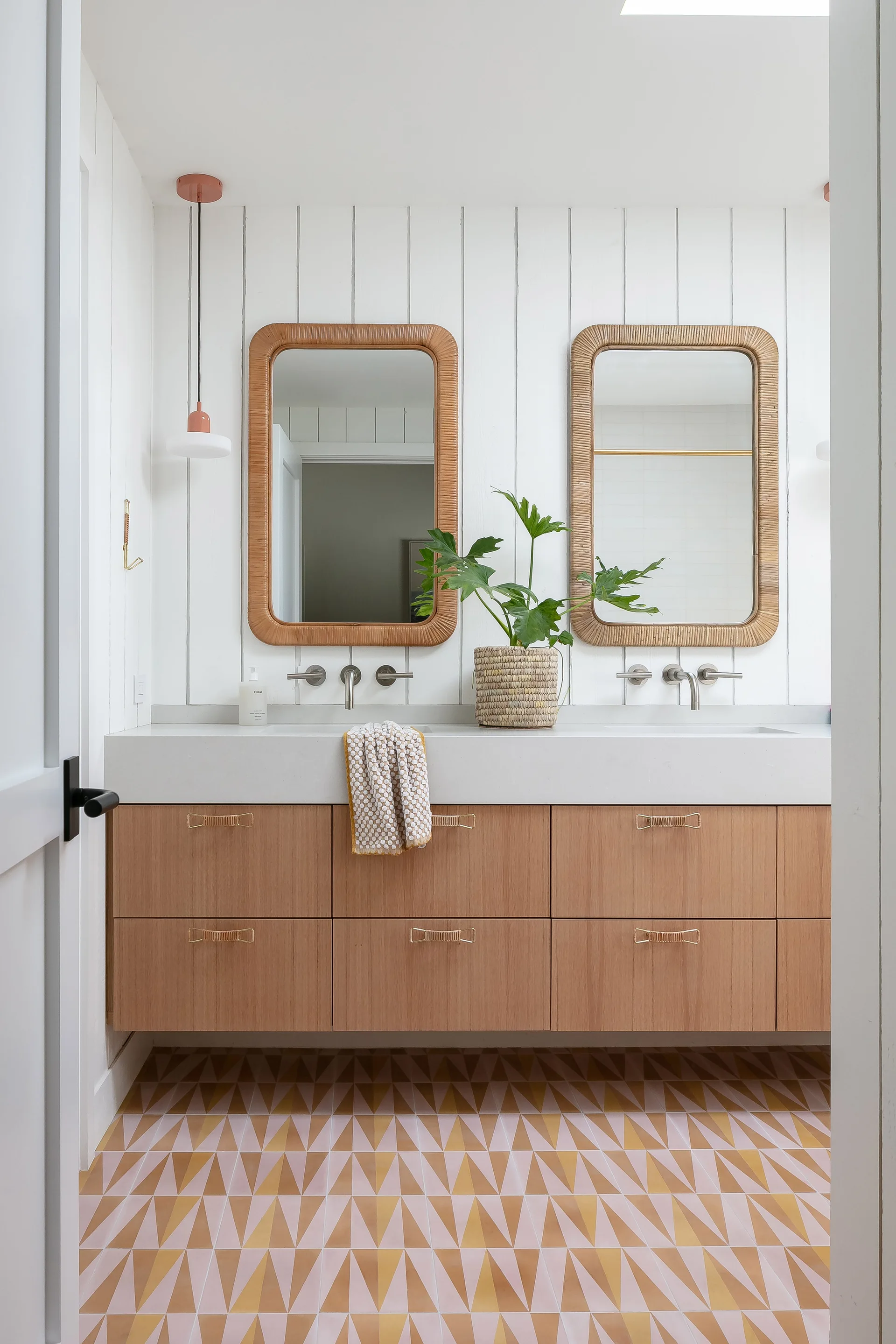 A modern bathroom features a floating wood vanity, dual rattan-framed mirrors, wall-mounted faucets, and a pink geometric patterned tile floor.