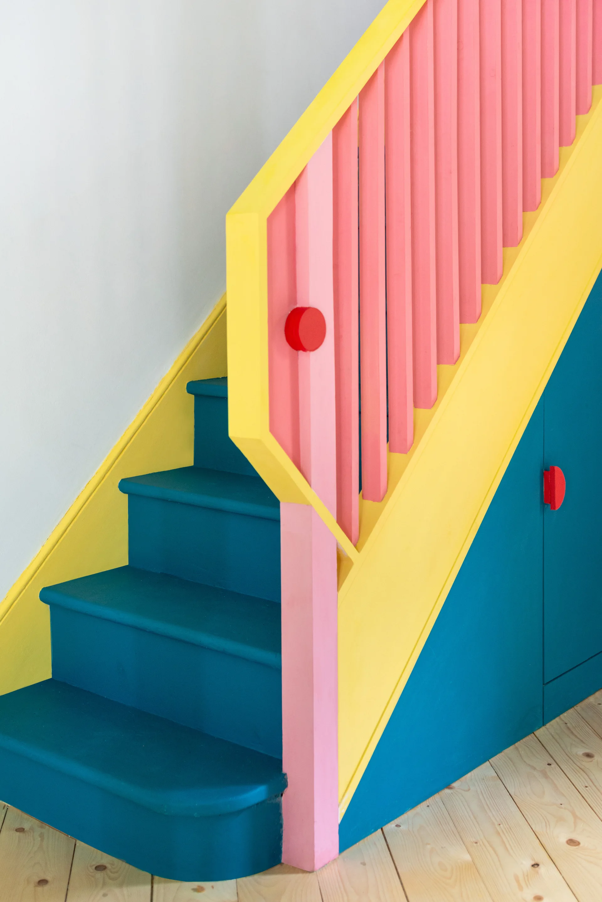 A colorful wooden staircase features blue steps, a yellow stringer, pink balusters, and a matching blue under-stair storage cabinet.