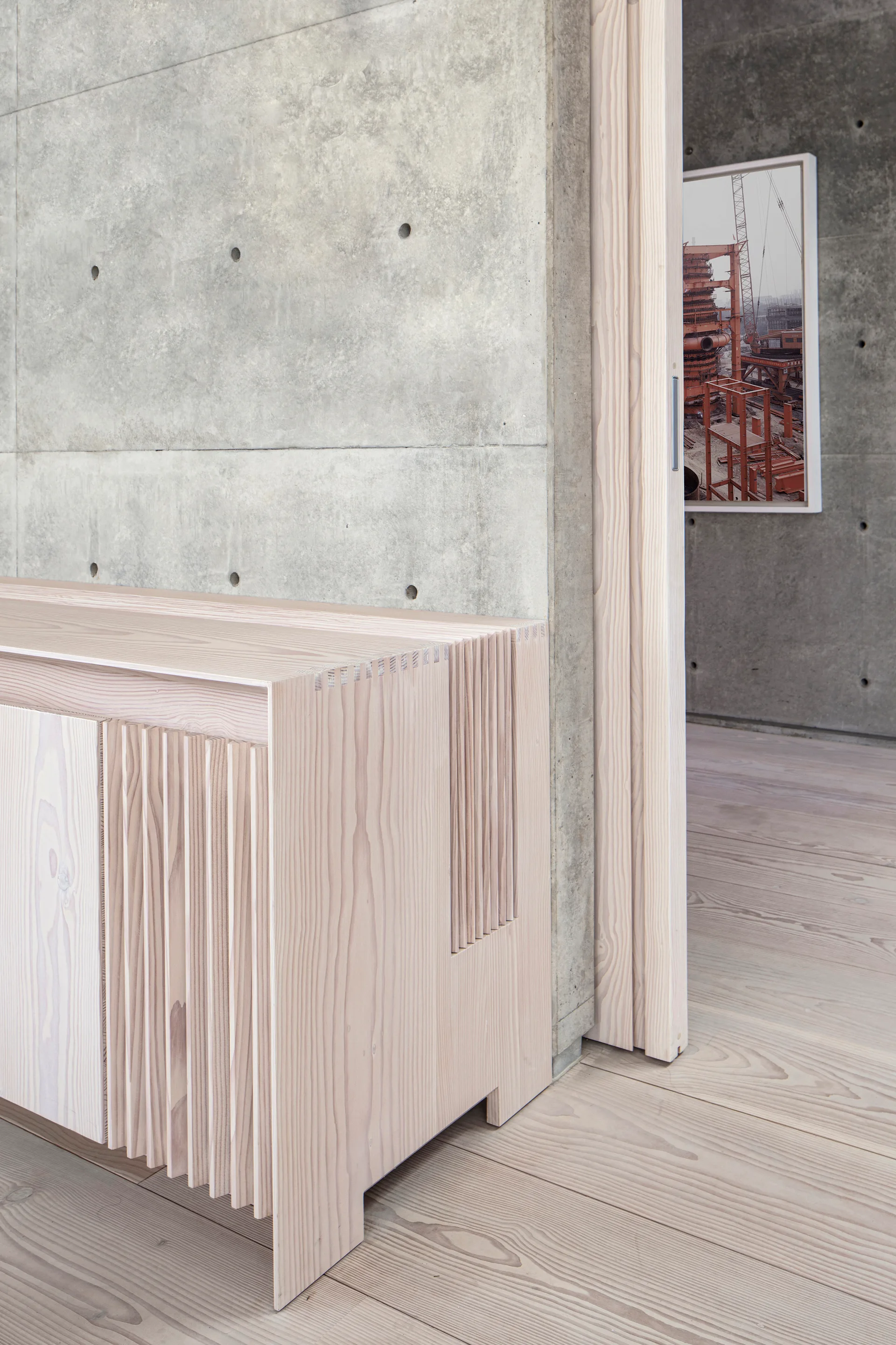 A pale wood console table with vertical slats sits against a grey concrete wall next to a sliding wood door.