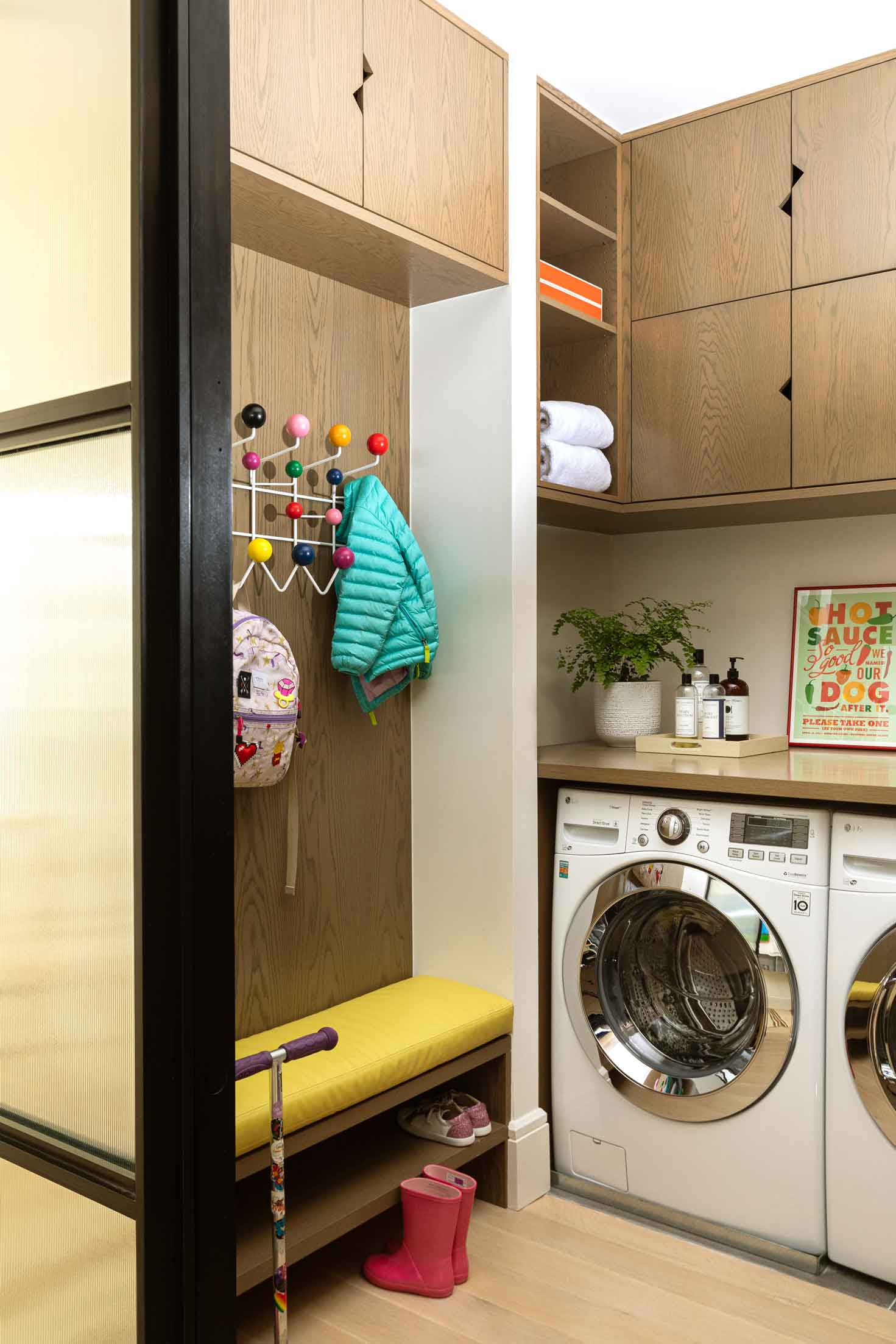 A laundry and mudroom space featuring light wood cabinets, a yellow cushioned bench, a colorful coat rack, and front loading washer.