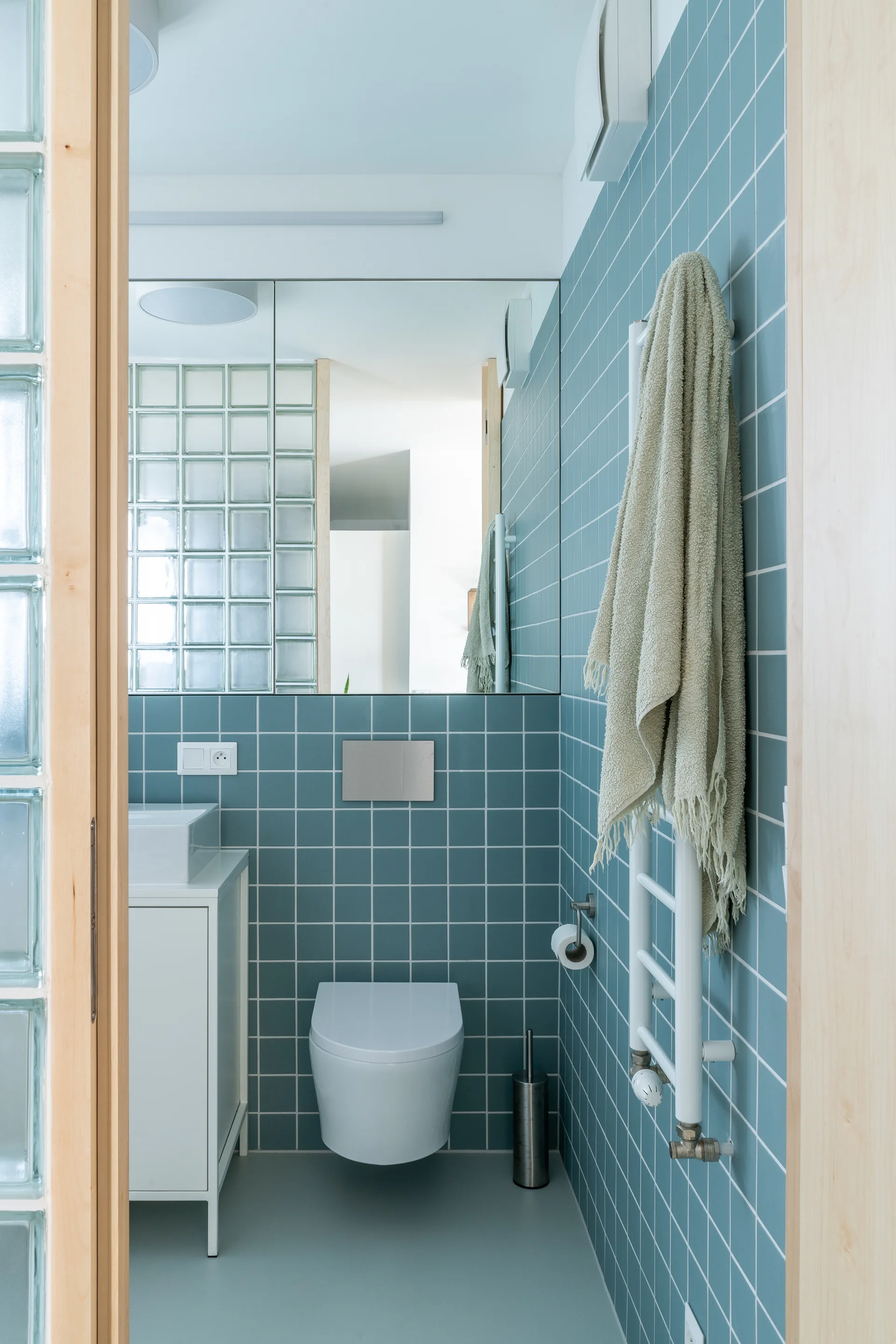 A modern bathroom features blue grid tiles on the wall, a white wall-mounted toilet, and a translucent glass block partition.