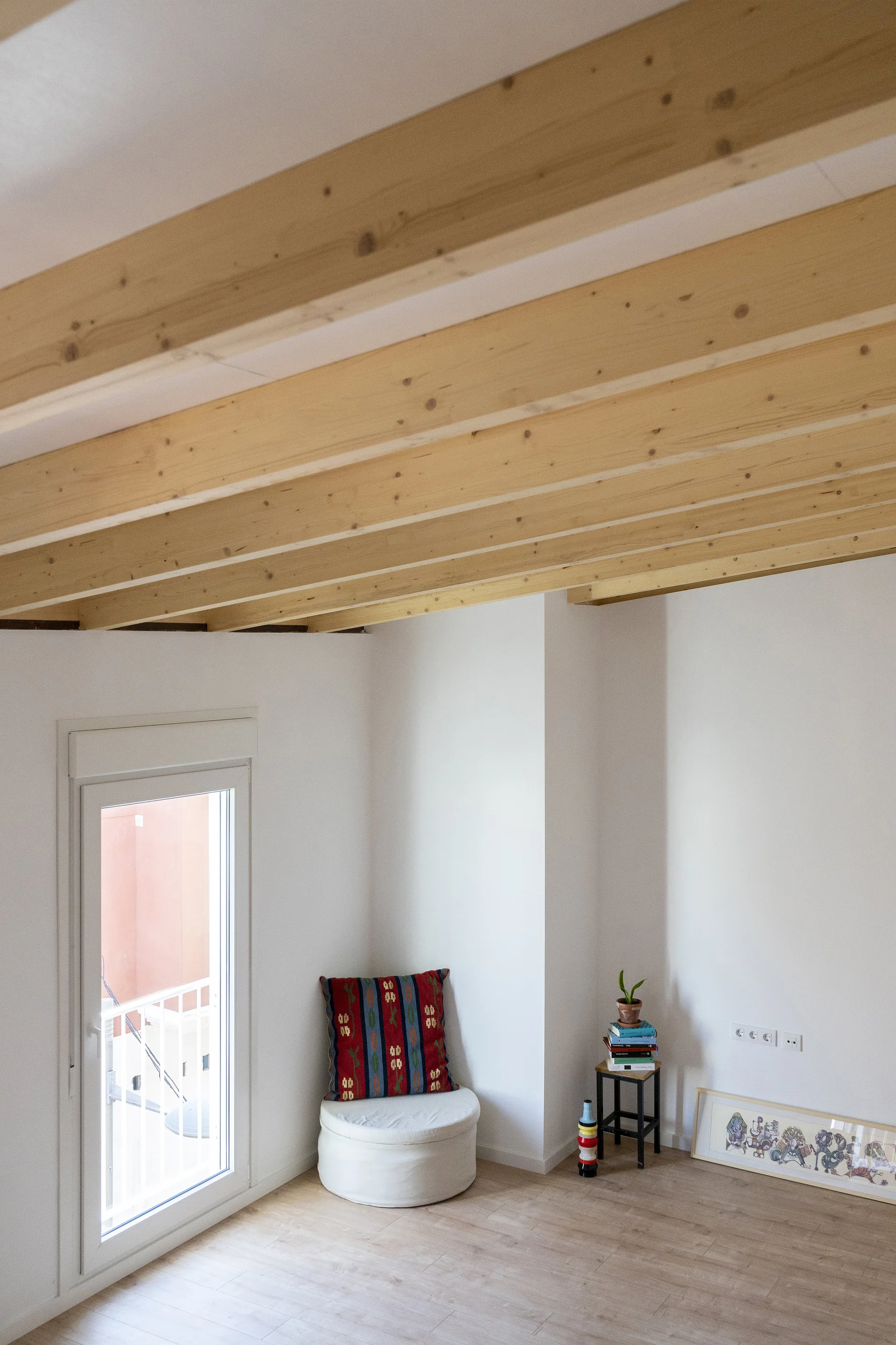 A minimalist room corner features exposed natural wood ceiling beams, white walls, and a low white ottoman with a patterned cushion.