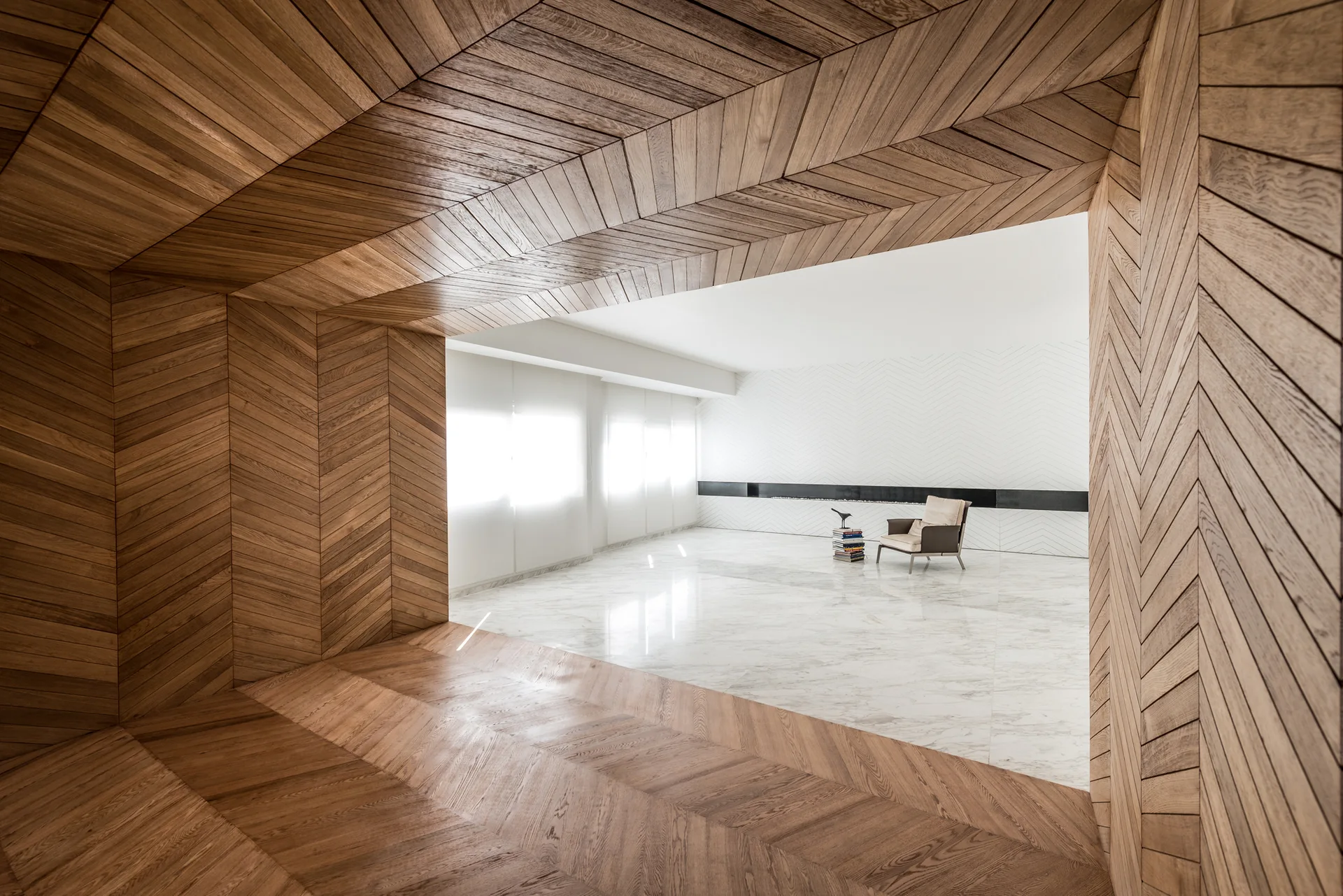 A minimalist living space featuring a faceted herringbone wood paneled entrance that opens into a bright room with polished white marble floors.