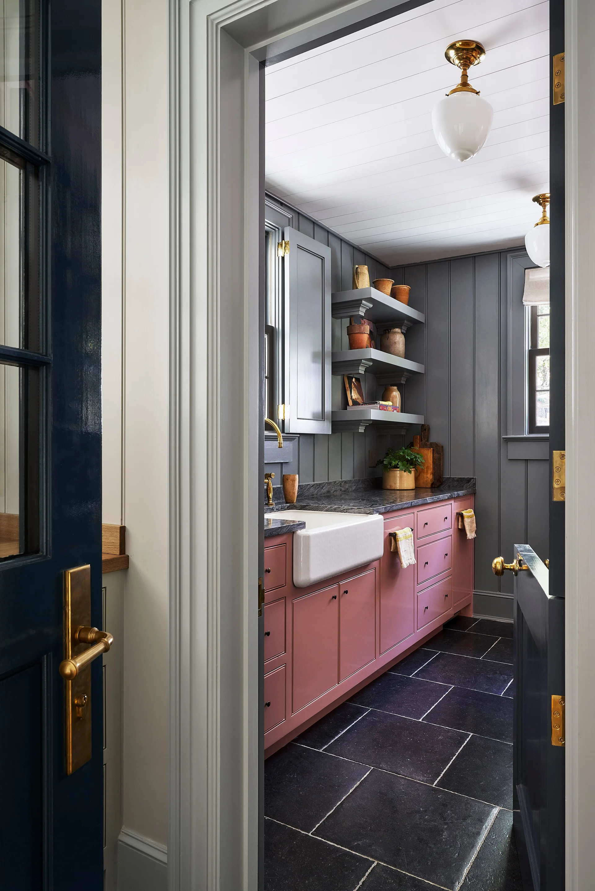 A view through a doorway into a kitchen featuring dusty pink cabinets, a white farmhouse sink, and gray vertically paneled walls.