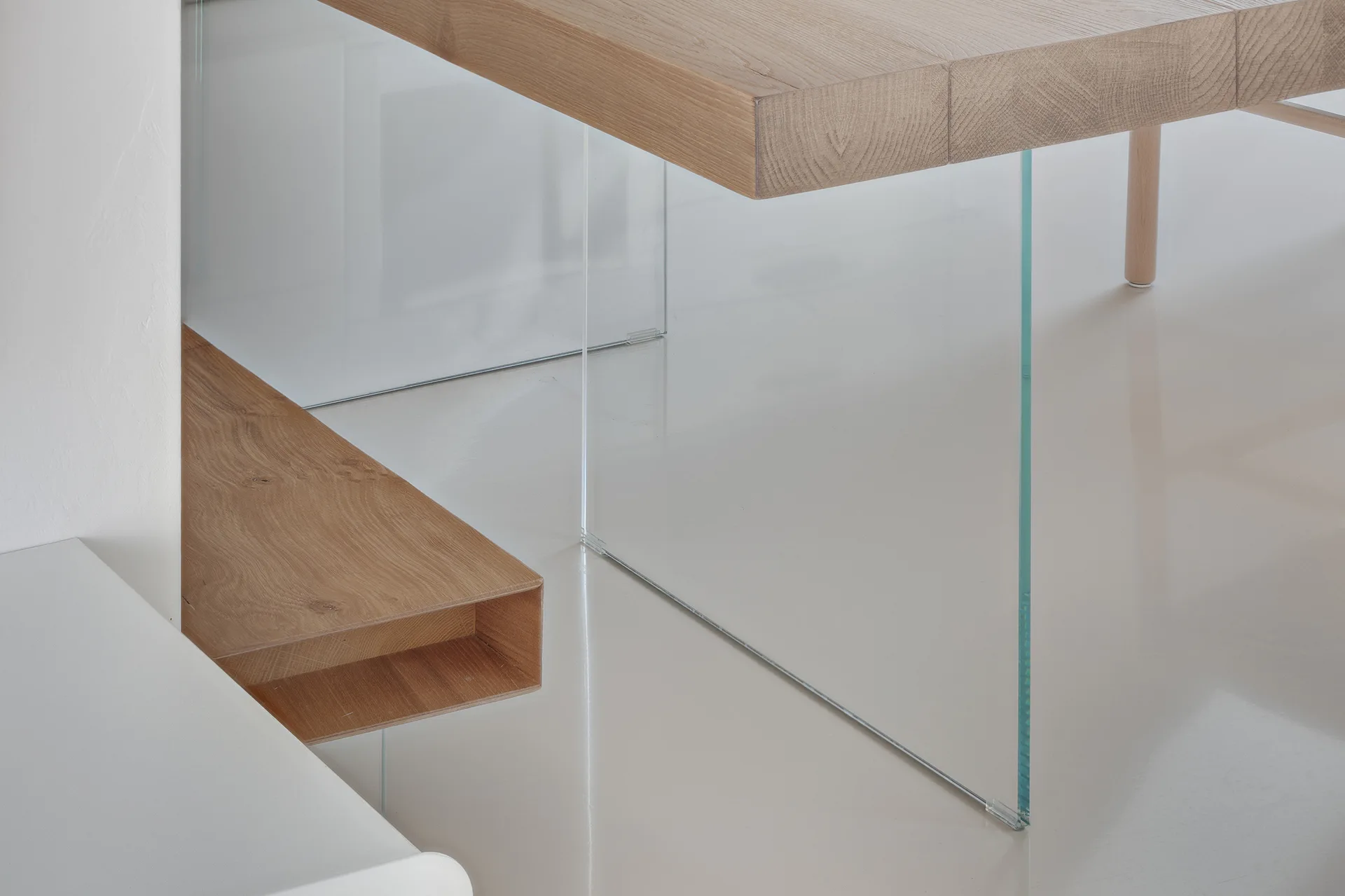 A close-up of a light oak dining table with a thick wood top supported by a transparent glass leg on a polished cream floor.