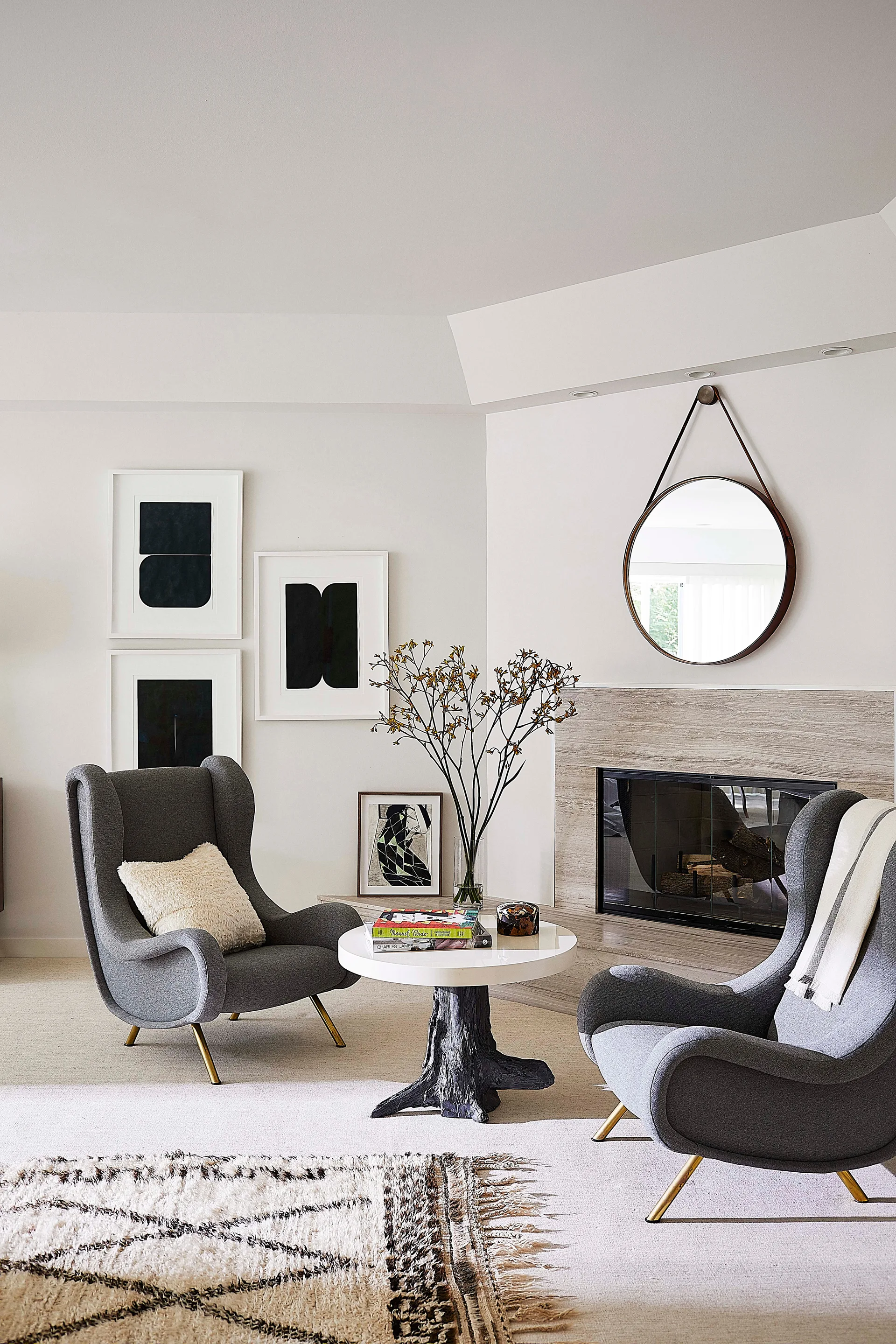 Two gray wingback armchairs face a round coffee table in a living room with a stone fireplace and suspended round mirror.