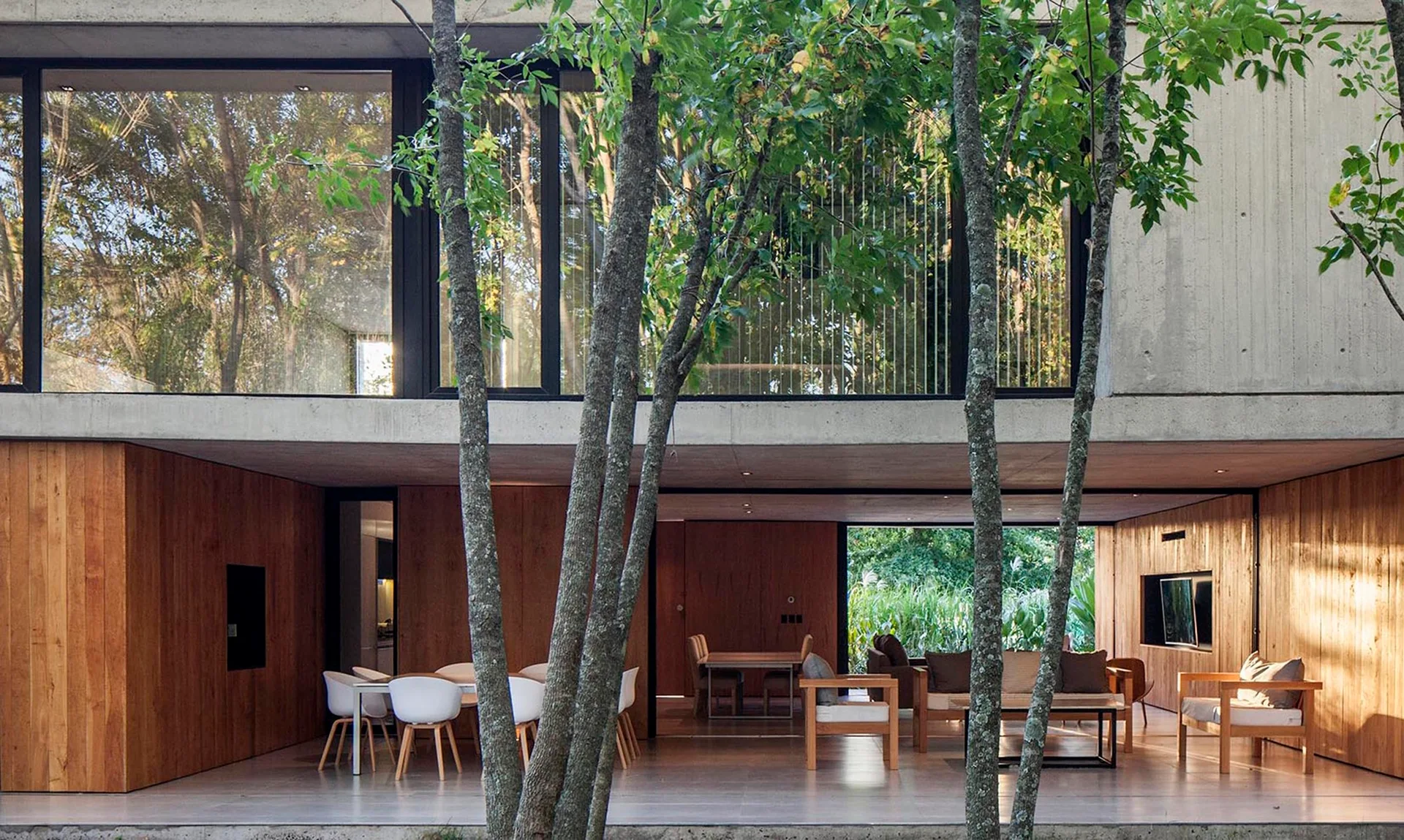 A modern open-concept home interior with warm wood-paneled walls, an exposed concrete ceiling, and large glass walls framing outdoor trees.