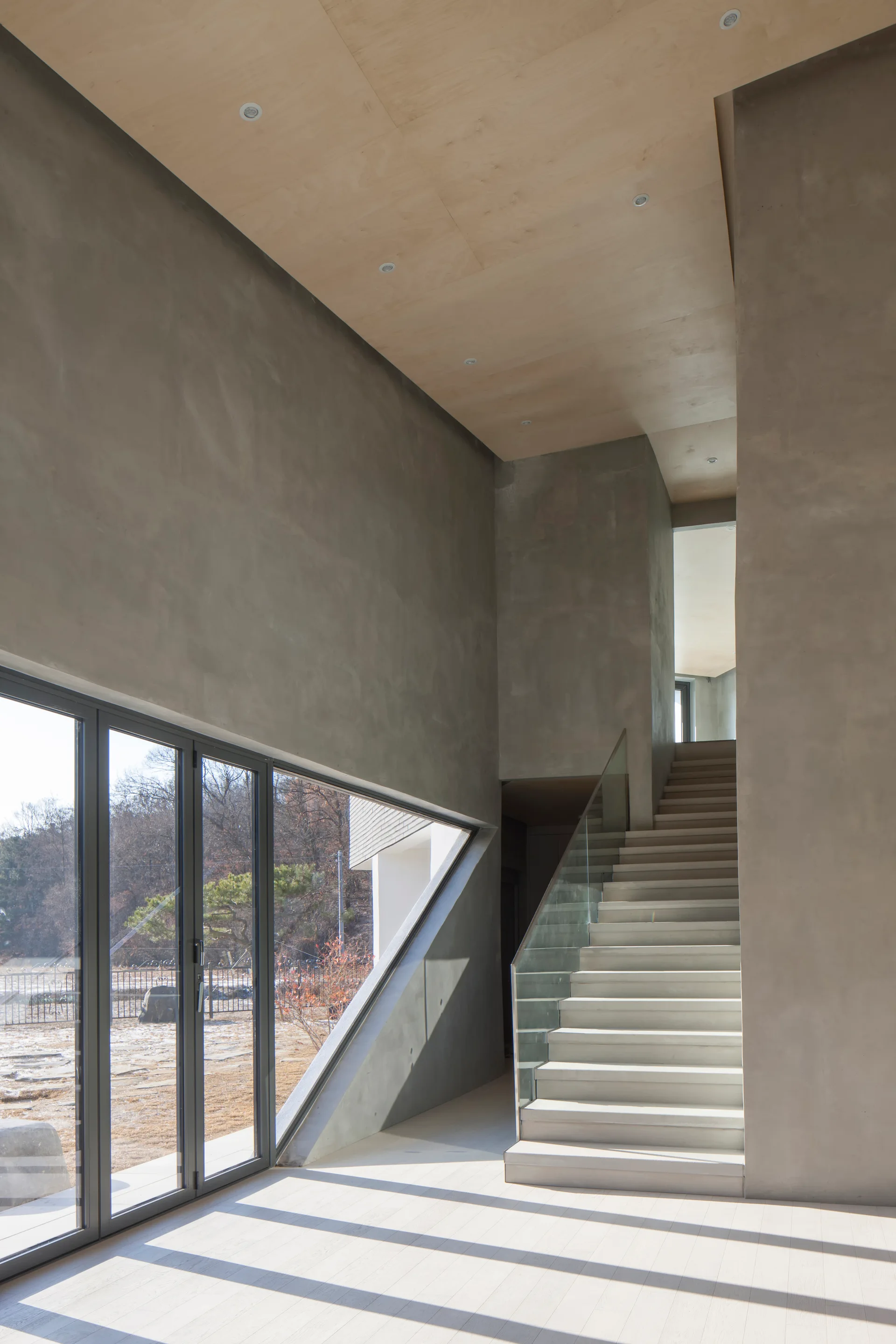 A modern interior features a concrete staircase with a glass railing, an angular window, concrete walls, and a light wood ceiling.